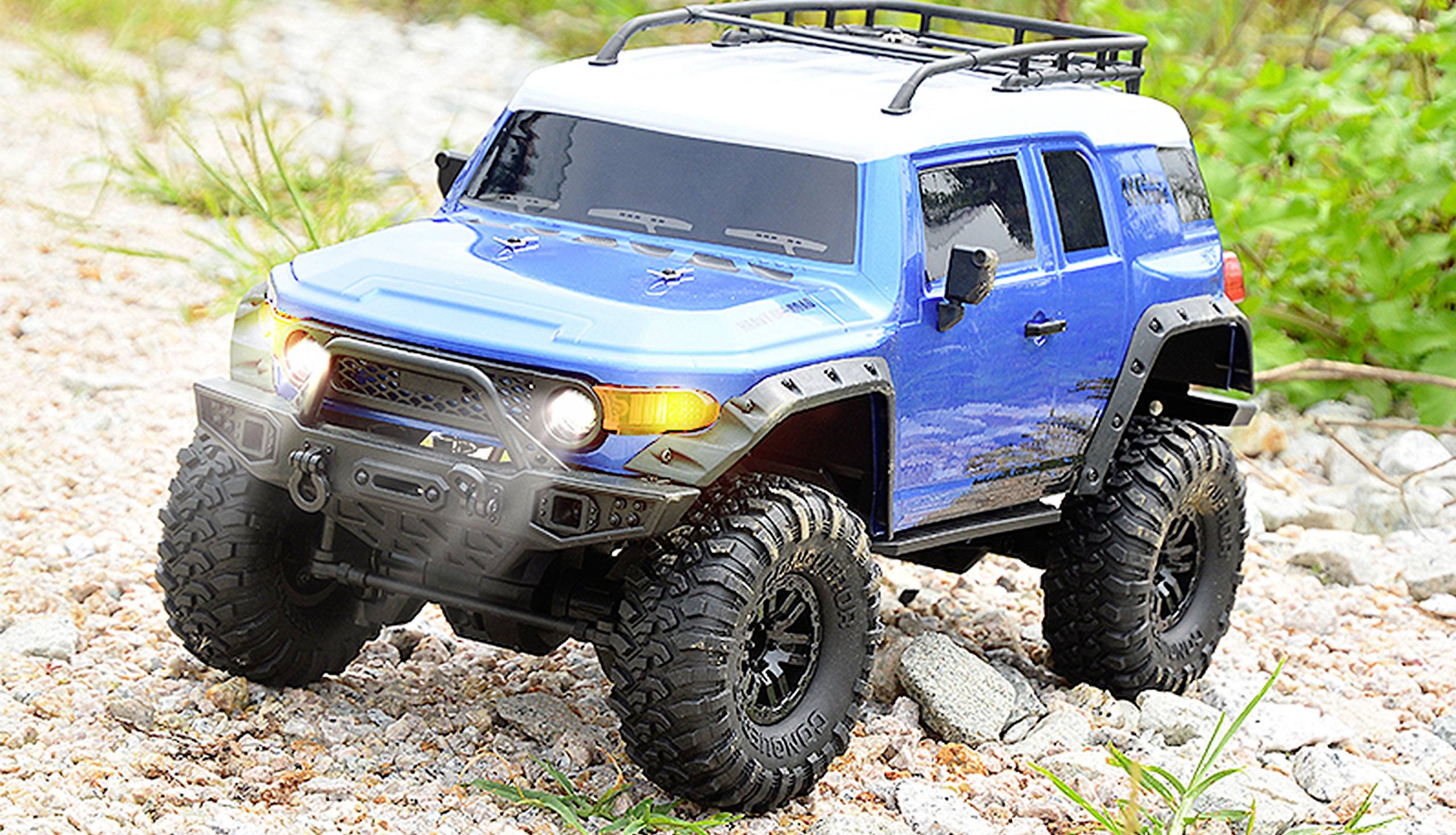 A blue remote-controlled off-road vehicle with large tyres drives over a gravelly surface, surrounded by green grass.