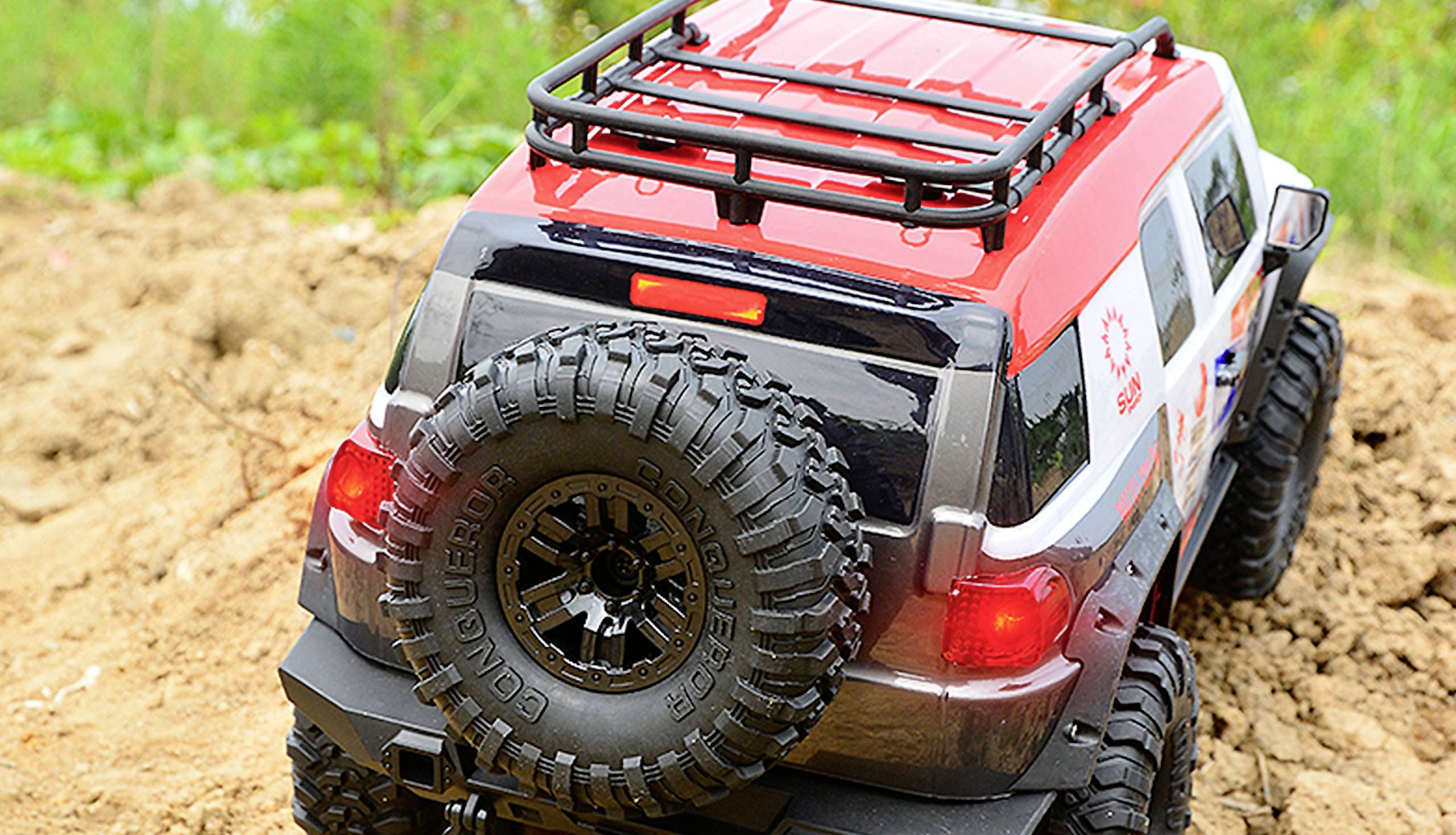 A red remote-controlled car drives over rough terrain. It has large tyres and a roof rack. The surroundings are earthy and grassy.