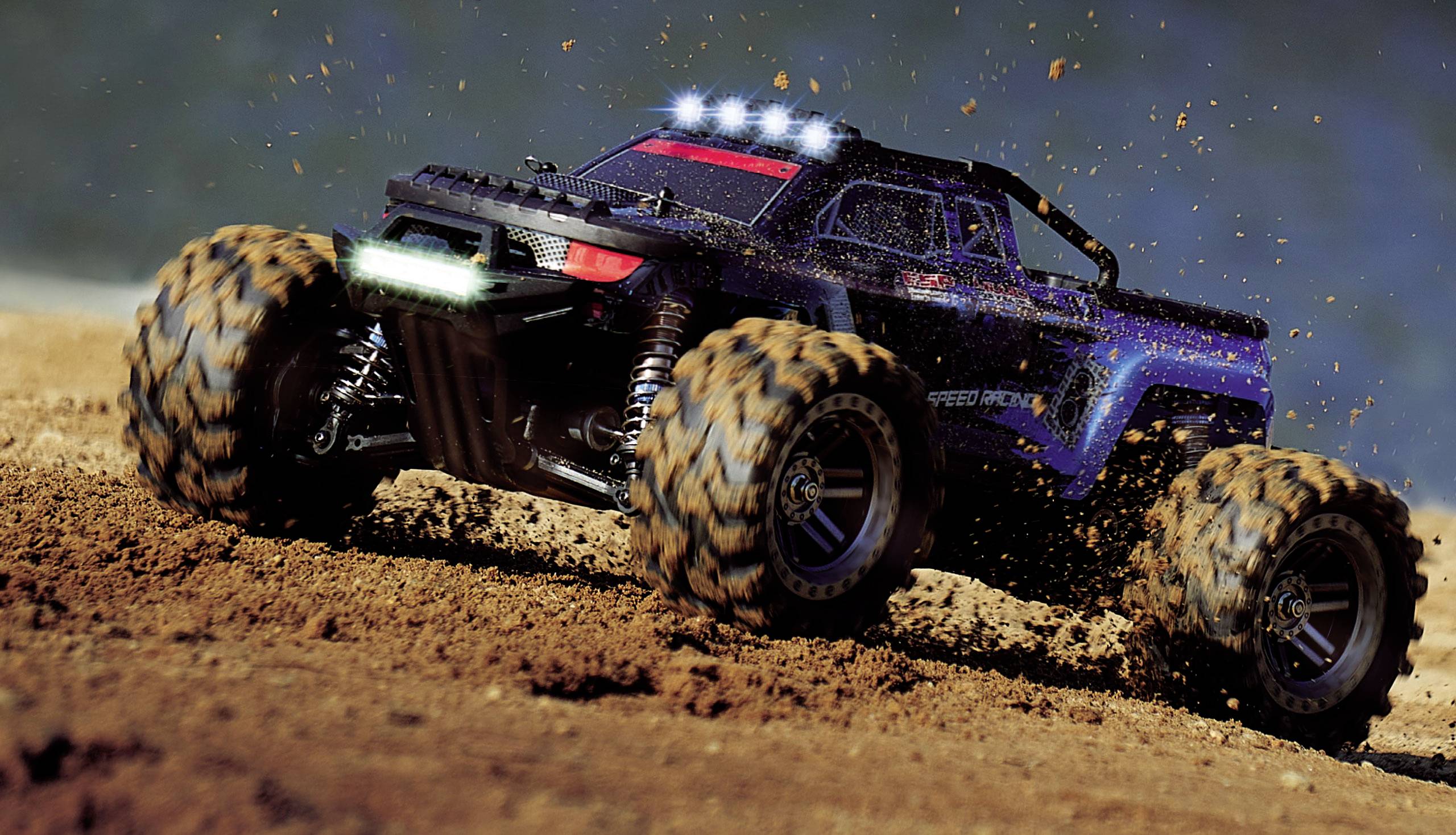 A remote-controlled car races across sandy ground at high speed. Dust and earth fly up behind its large tyres.
