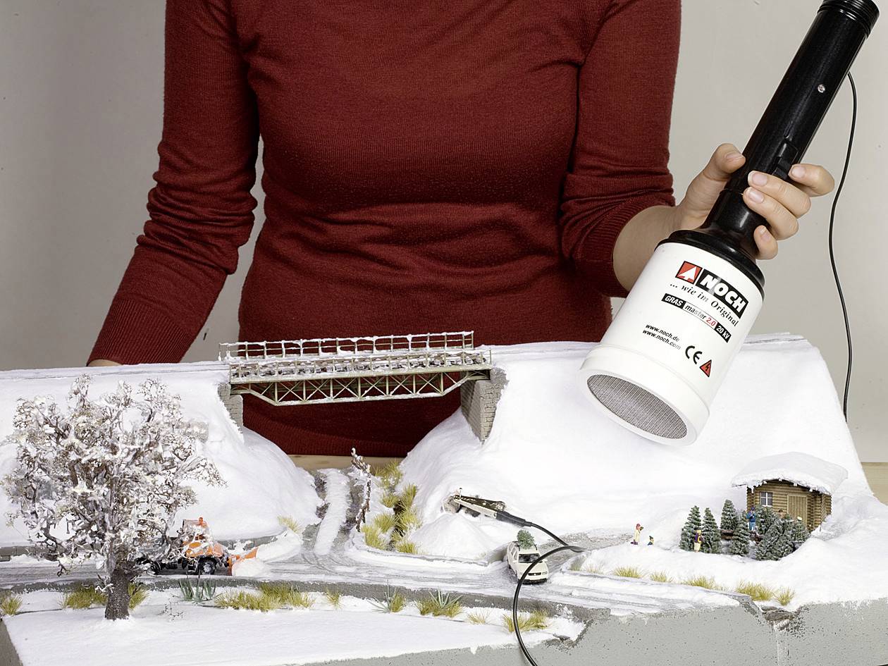 A person illuminates a model of a wintry landscape with a bridge, roads, and trees. Snow and vehicles are in the foreground.