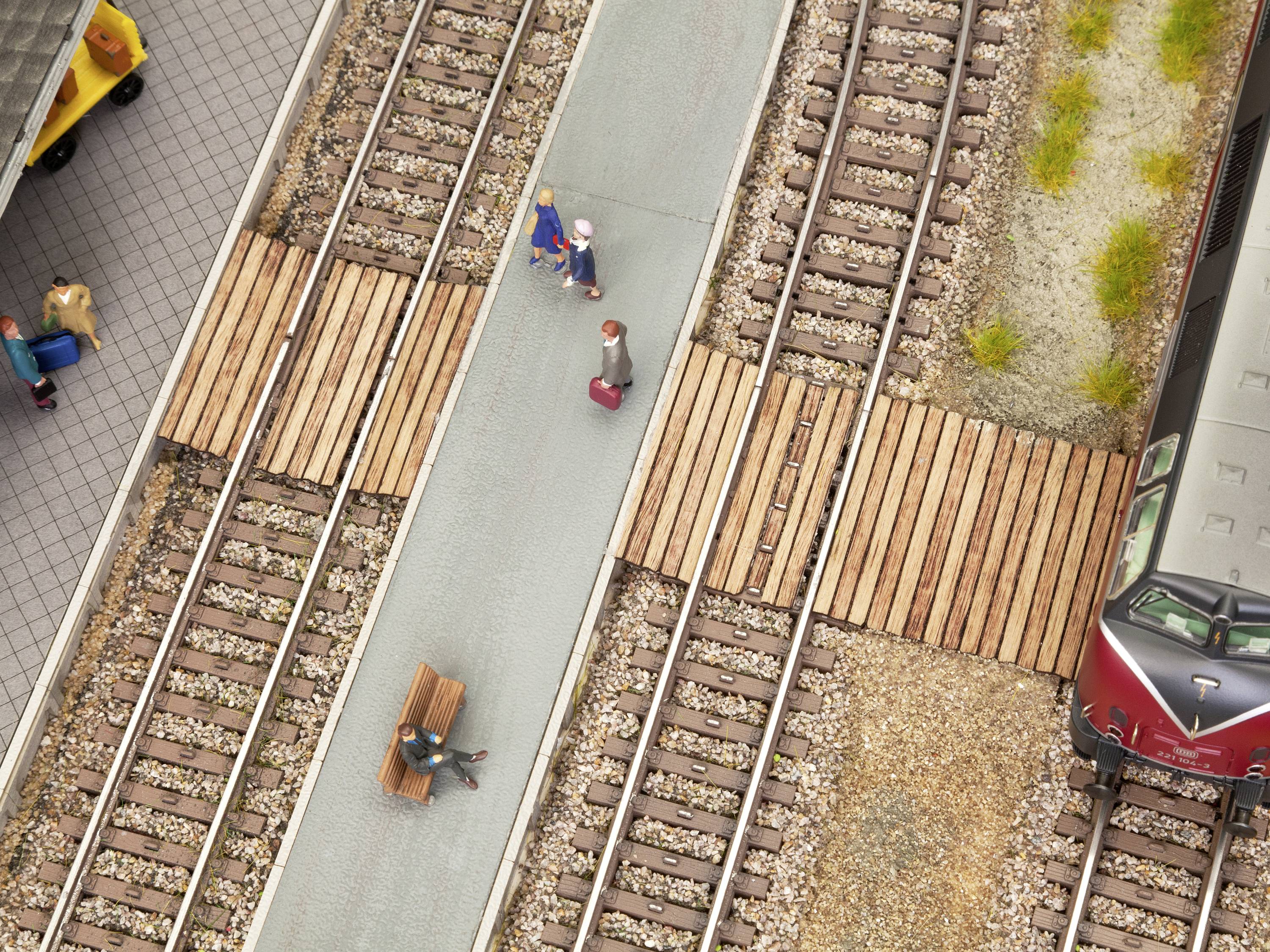 View of a railway station with people walking across a platform. Several tracks run parallel, with a train stationed on the right.