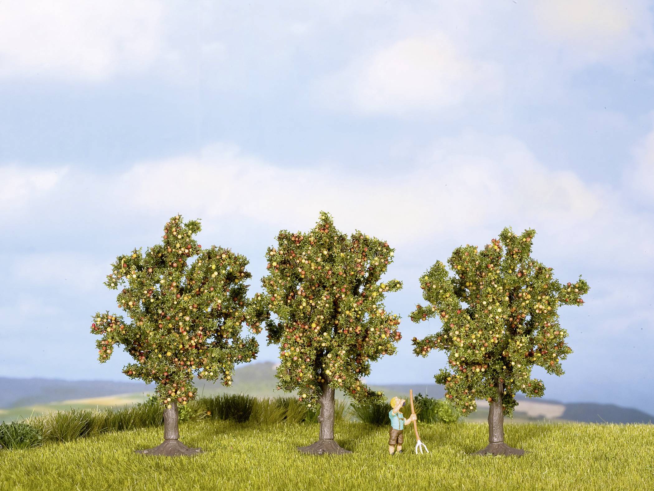 Three apple trees on a meadow, with a small model of a person holding a ladder. The sky is clear and blue.