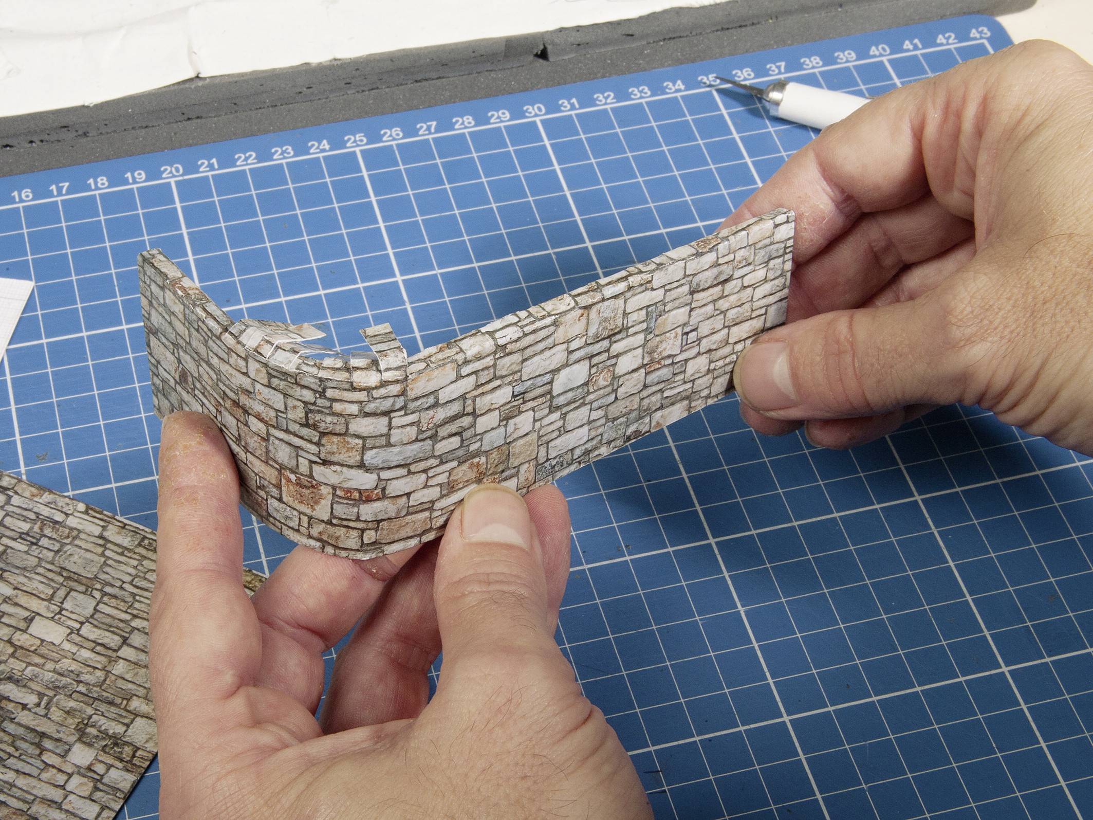 Two hands are holding a piece of model wall on a blue cutting board, surrounded by craft materials.