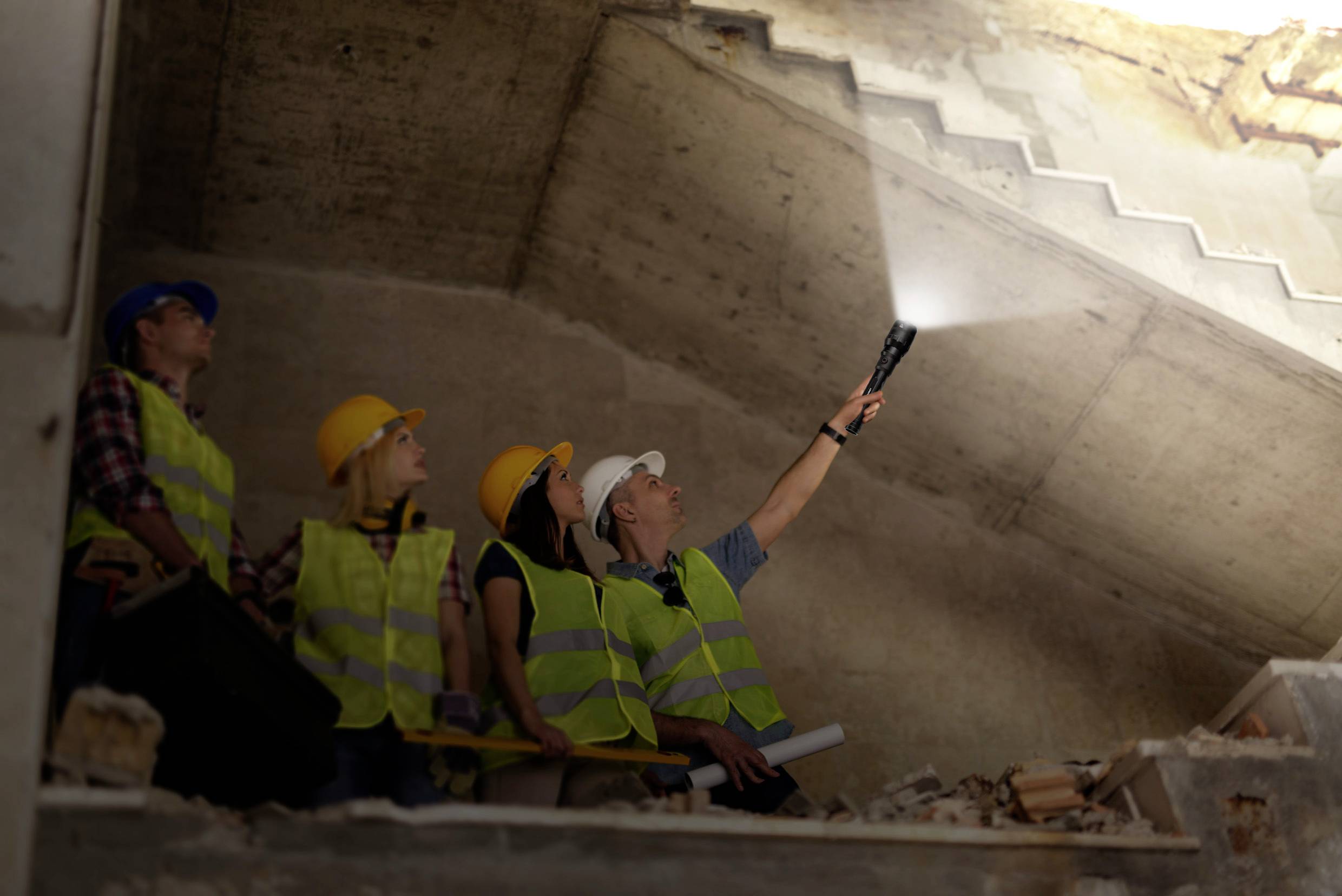 Workers inspect a construction site in an unfinished building. One person is shining a torch on the ceiling. Everyone is wearing hard hats and high-visibility vests.