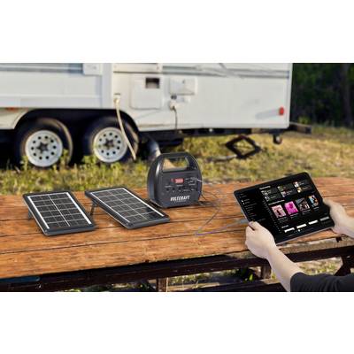 Solar panels and charger on a wooden table, connected to a tablet, with a caravan in the background. Charging on the go.