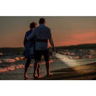 An elderly couple walks along the beach at sunset. The man is holding a small glowing lantern in his hand.