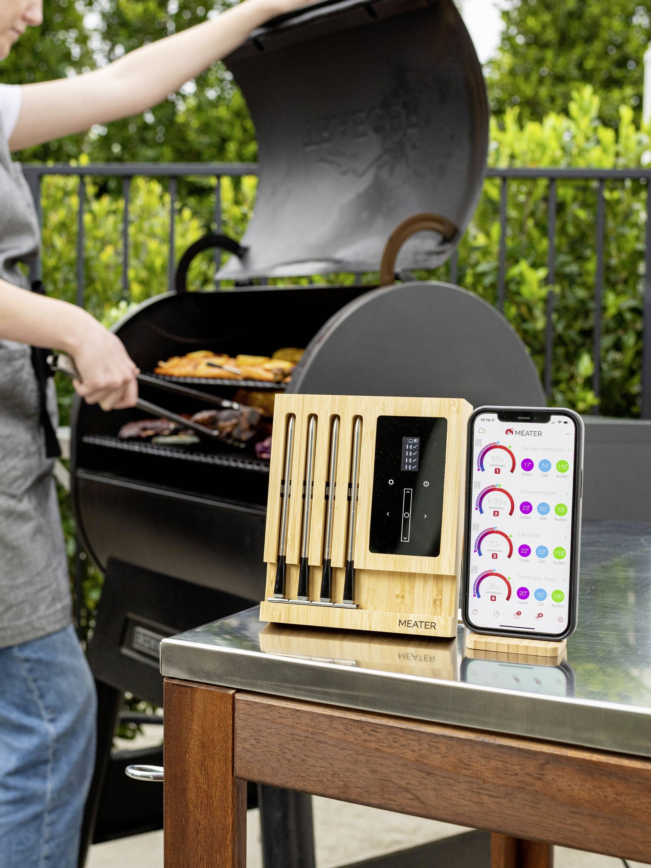 A person is grilling meat on an outdoor barbecue. In the foreground, there is a meat thermometer set and a smartphone with an app display.