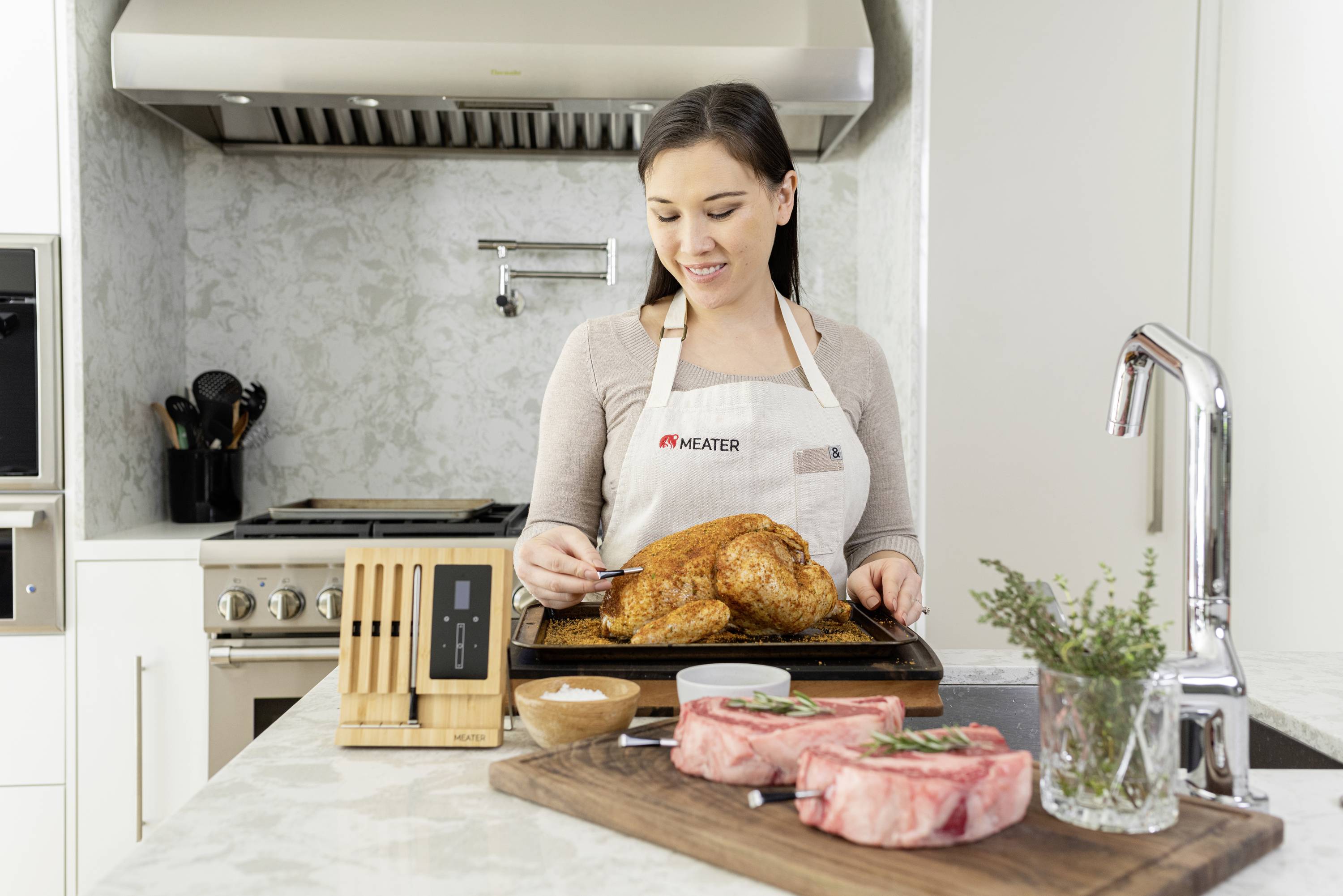 A woman in a modern kitchen is checking the temperature of a roasted chicken with a meat thermometer. Two raw pieces of meat lie in front of her.