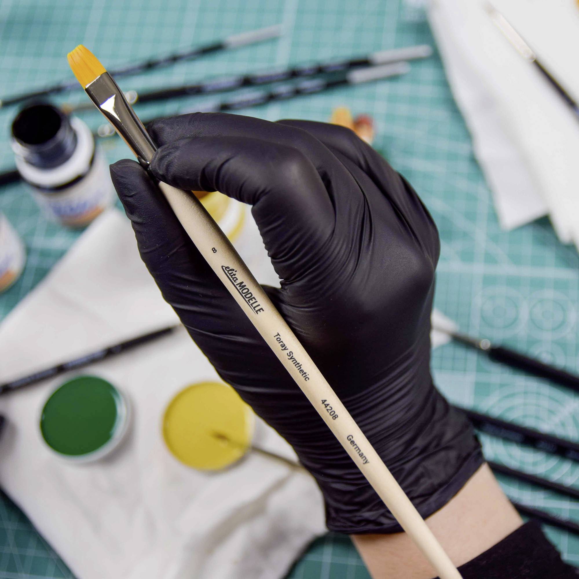 A hand wearing a black glove is holding a paintbrush over a table with paints and art materials.