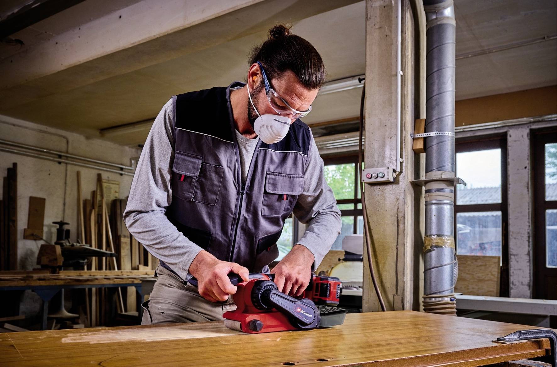 A person is sanding wood with an electric sander in a workshop. They are wearing safety glasses and a dust mask.