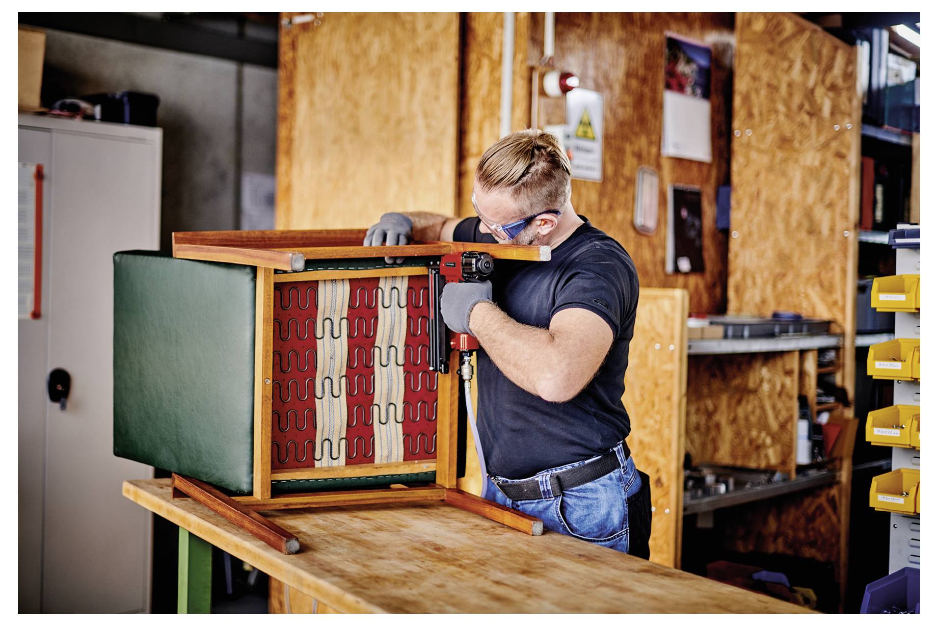Man wearing gloves and safety goggles assembles a wooden cabinet or furniture frame in a workshop.