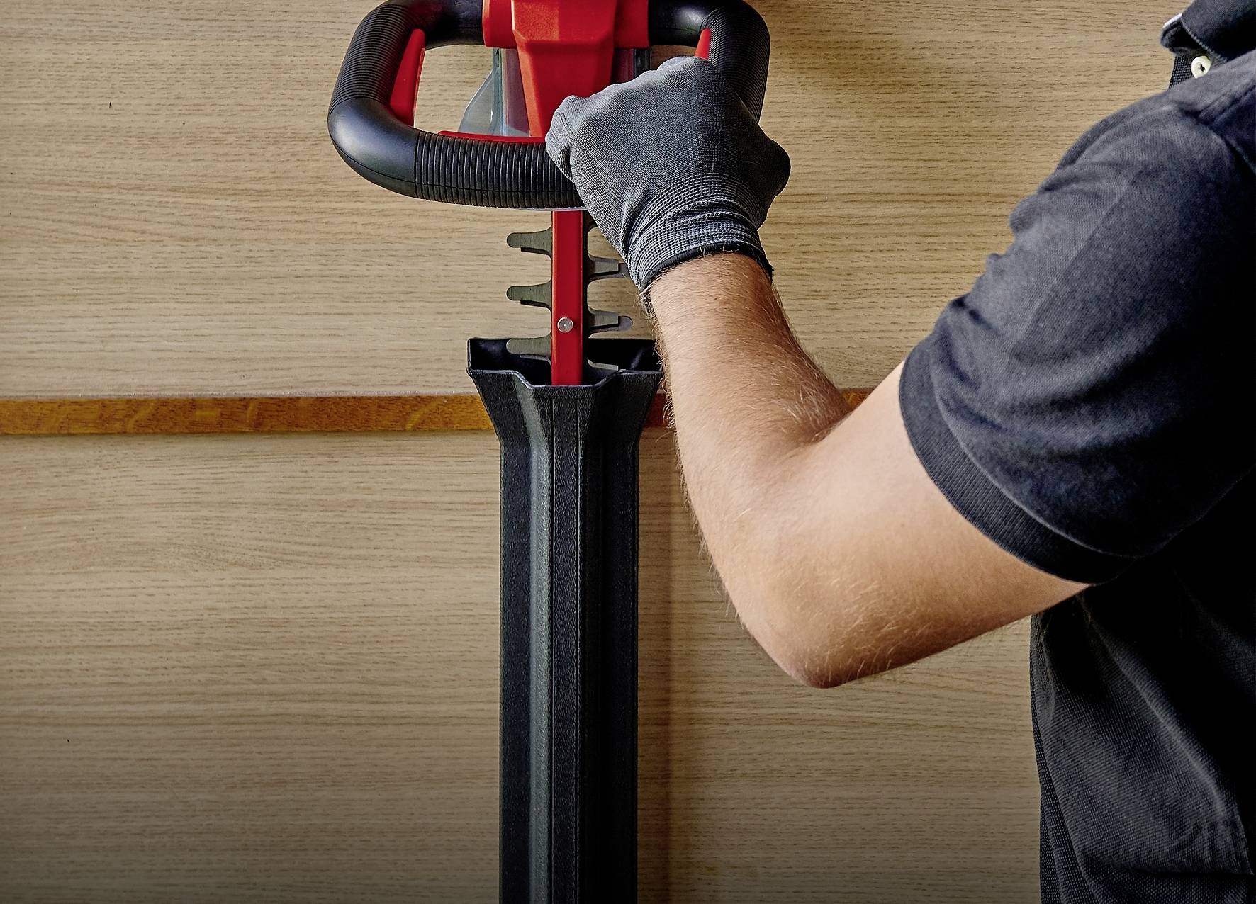 A person wearing a black shirt and gloves is operating a machine with red and black components in front of a wooden wall.
