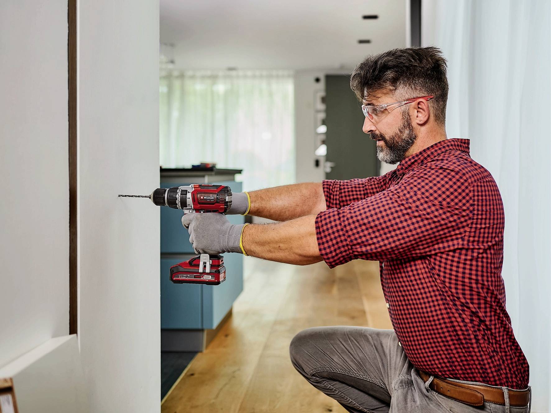 A man in a checked shirt is using a drill to work in a modern apartment. He is wearing safety glasses and gloves.
