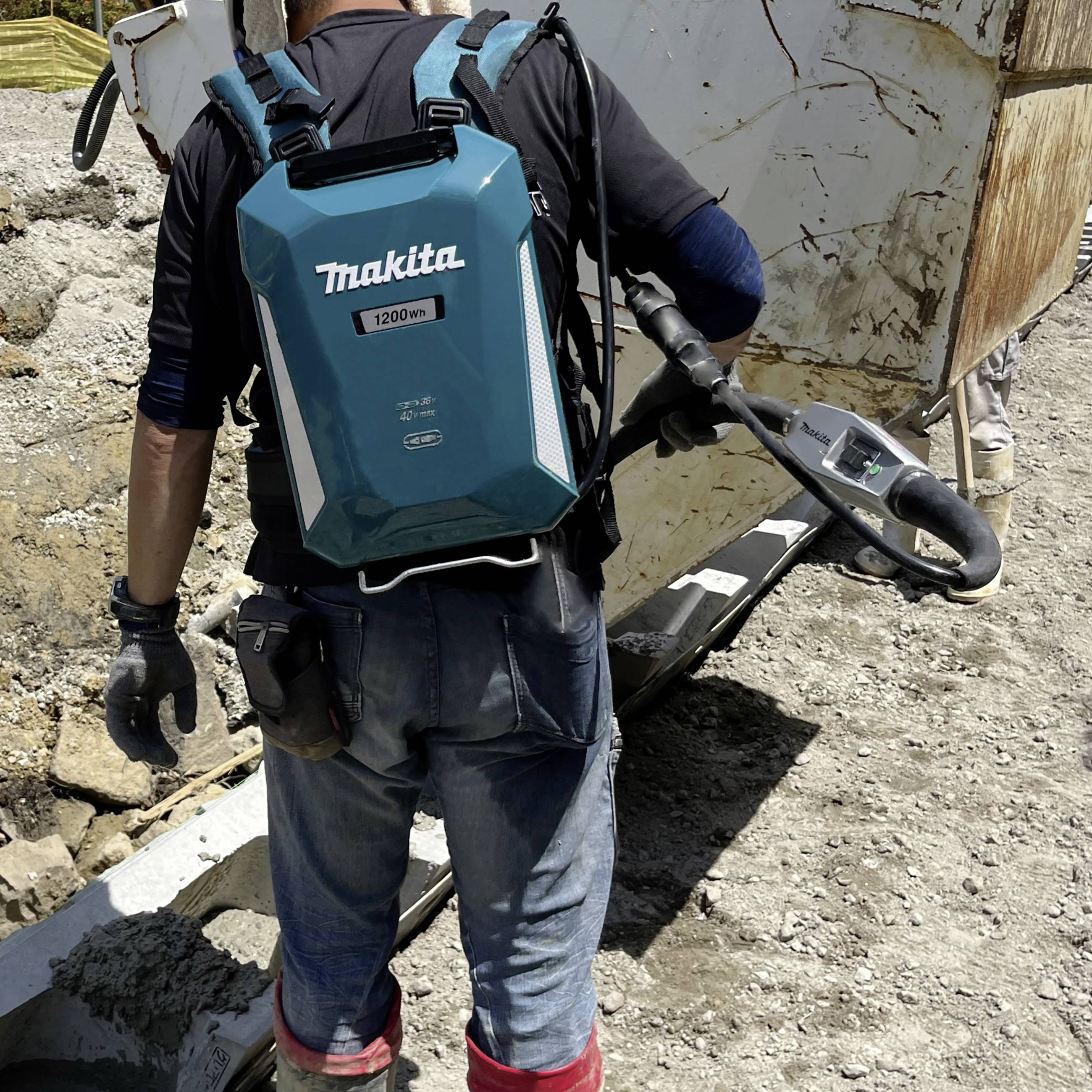 A person on a building site is wearing a large Makita backpack and manoeuvring a heavy, mobile piece of equipment across uneven ground.