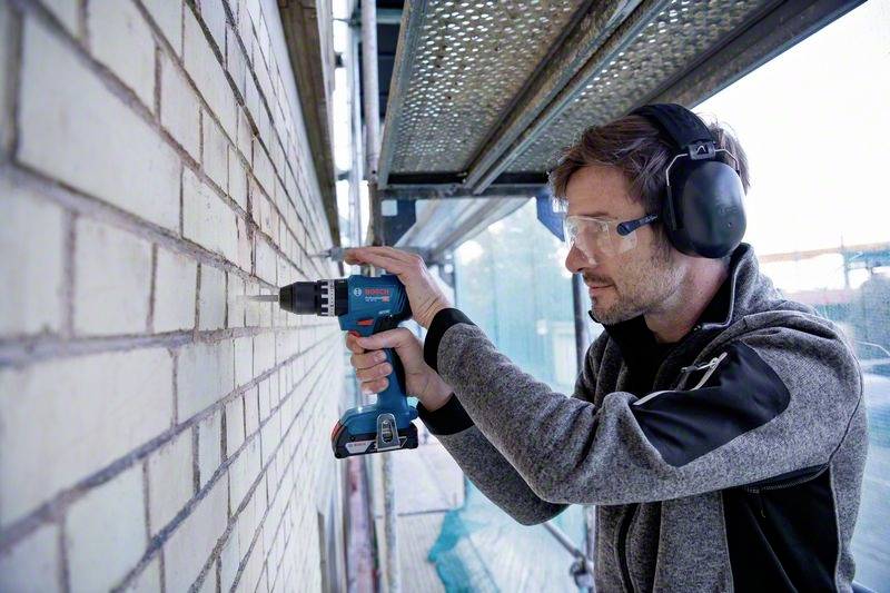 A person is drilling holes in a brick wall using a cordless drill. They are wearing ear protection and safety glasses, and are standing on a scaffold.