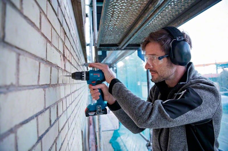 A person is drilling holes in a brick wall using a cordless drill. They are wearing ear protection and safety glasses, and are standing on a scaffold.
