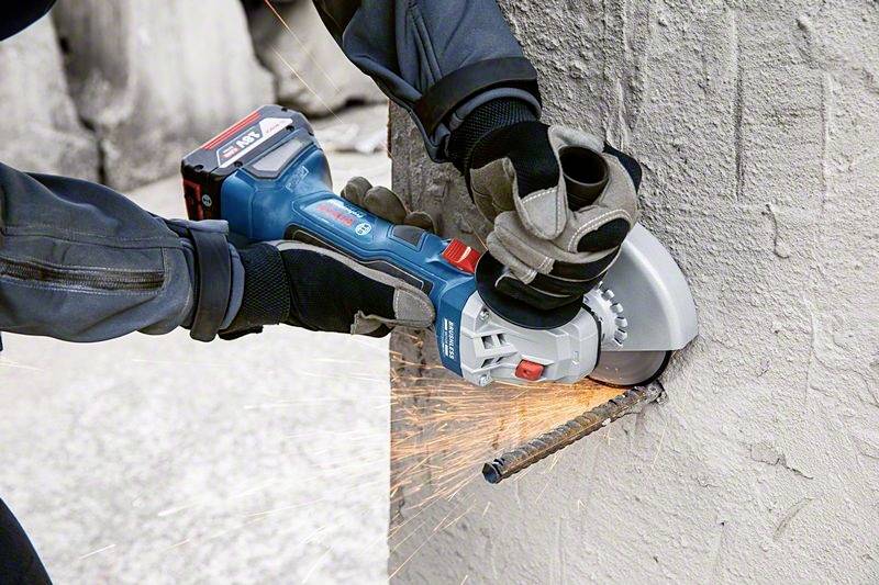 A person is cutting a metal wire from a concrete wall using a cordless angle grinder, with sparks flying.