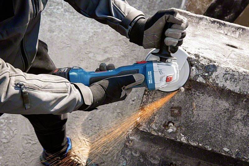 A person is wearing protective clothing and using an angle grinder to cut metal. Sparks are flying from the device.
