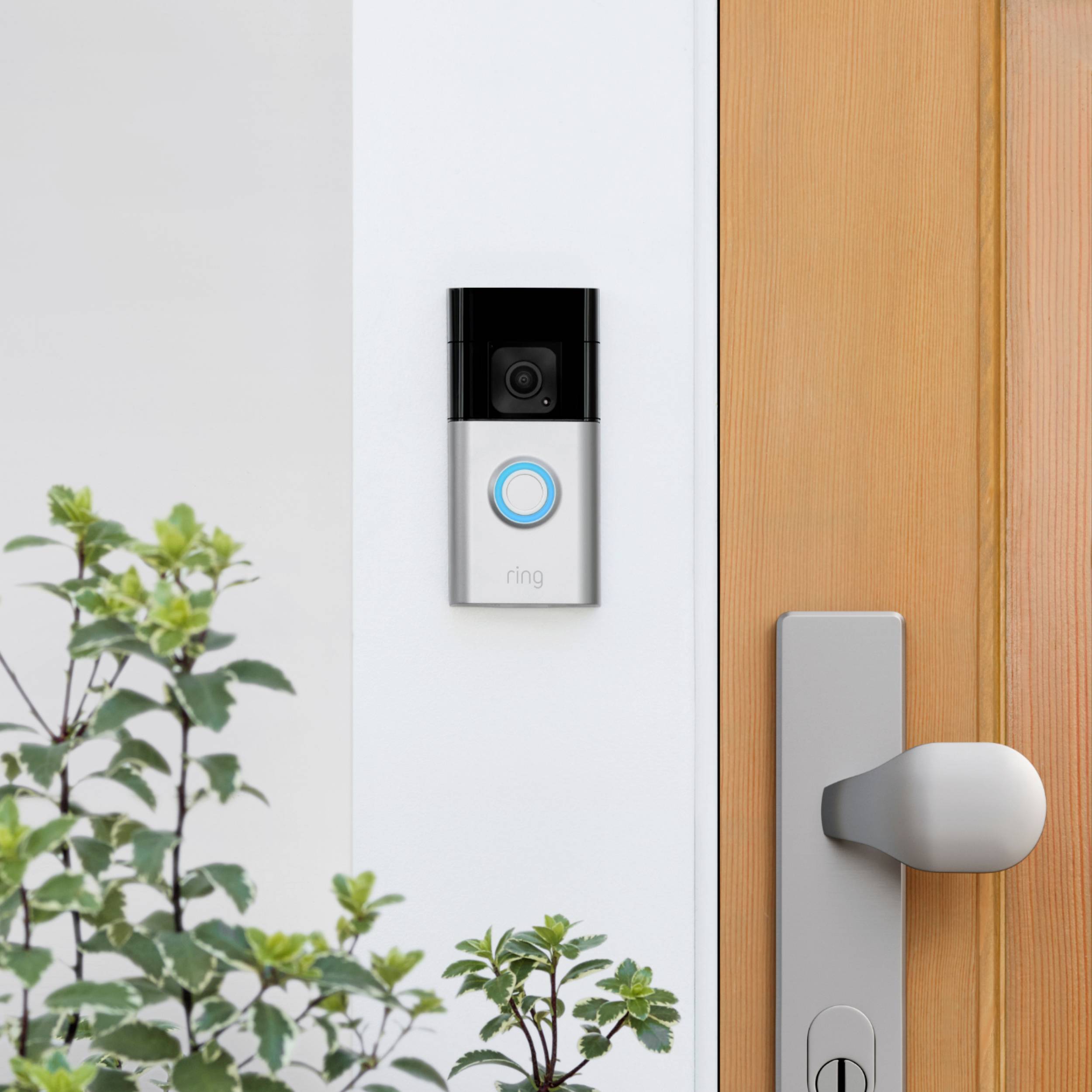A video doorbell is installed on a white wall beside a wooden door, surrounded by green plants.