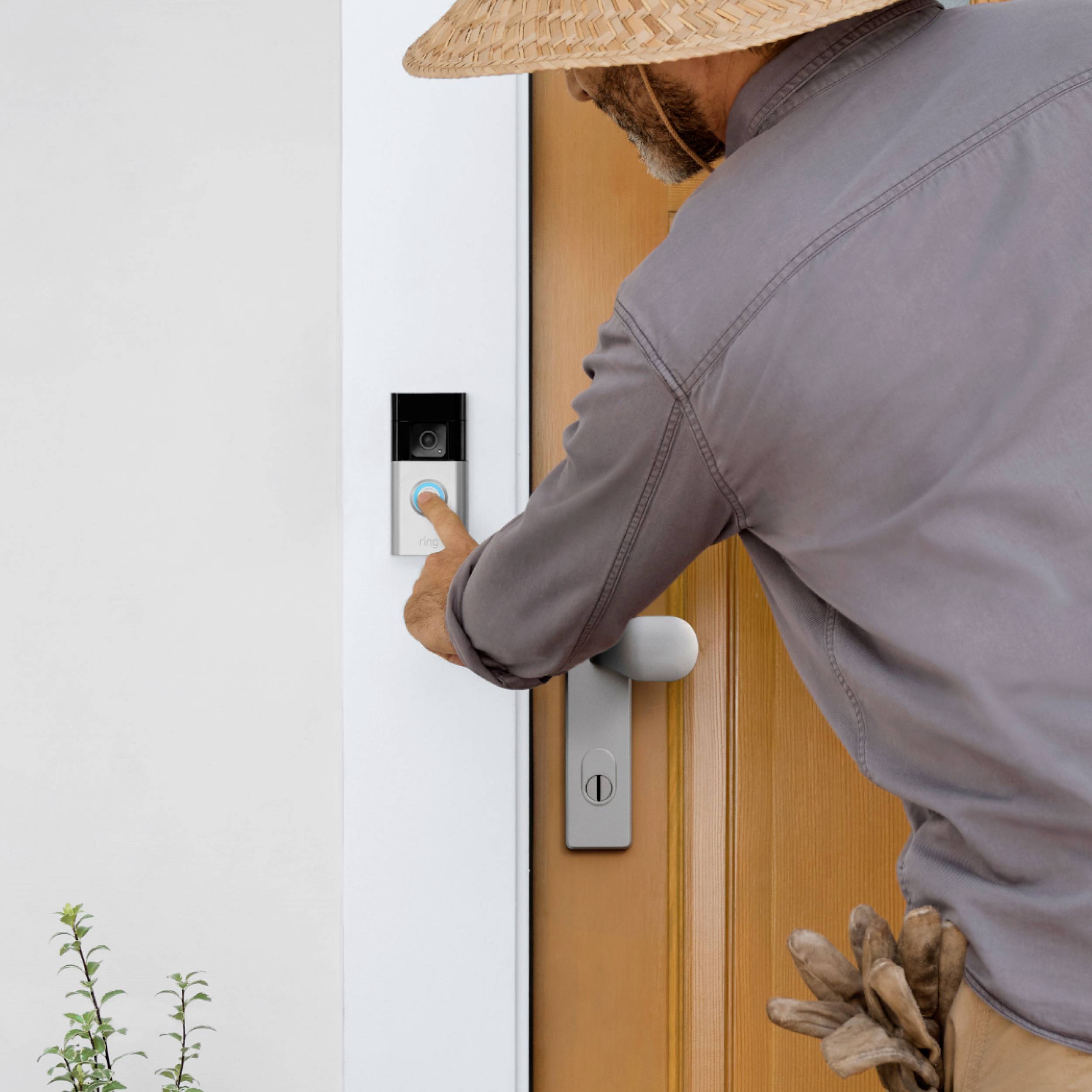A person presses the doorbell on a door with a video camera installed. They are wearing a straw hat.