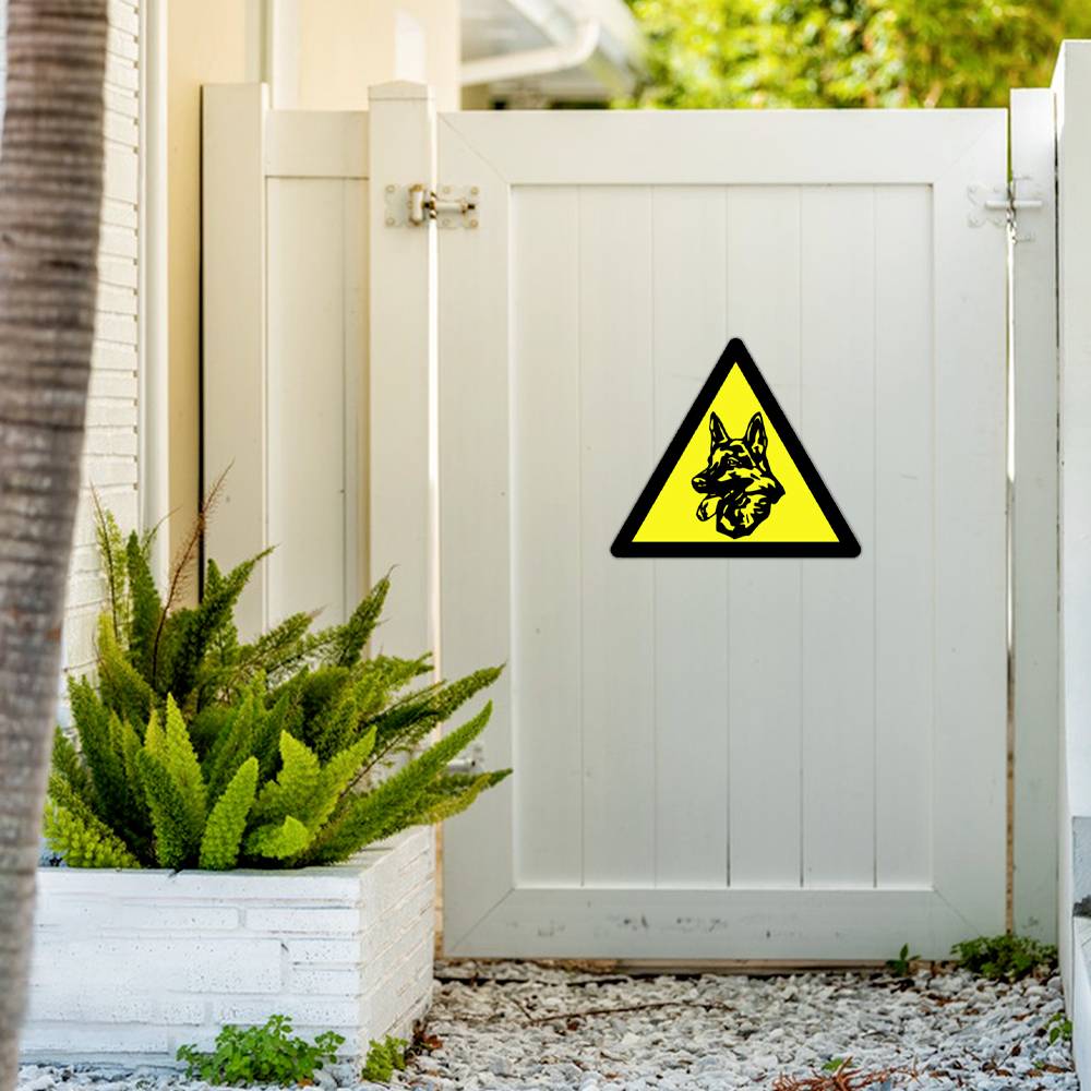 A white gate with a yellow warning sign displays the silhouette of a dog's head. Plants stand to the left of the gate.