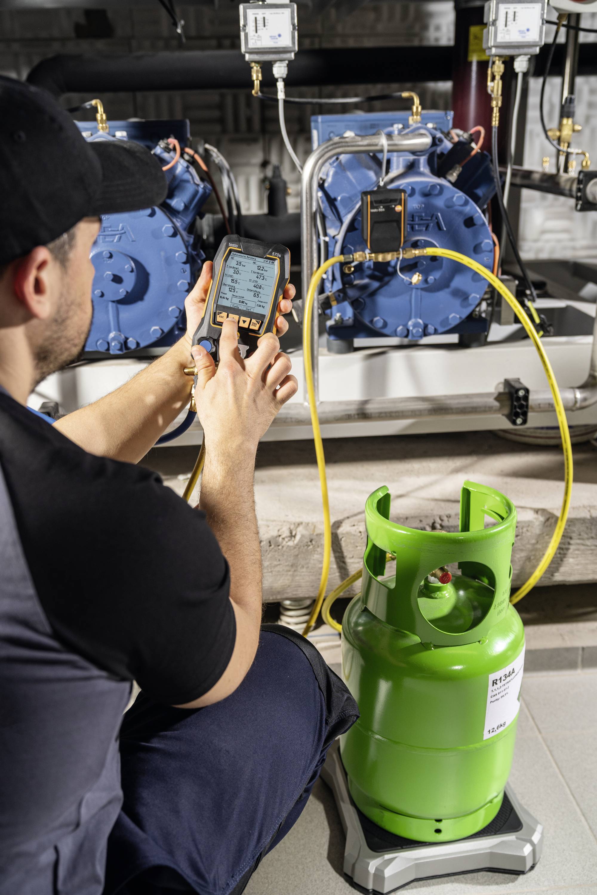 A technician checks the pressure of refrigeration compressors using a device. A green gas cylinder stands on a scale in the foreground.