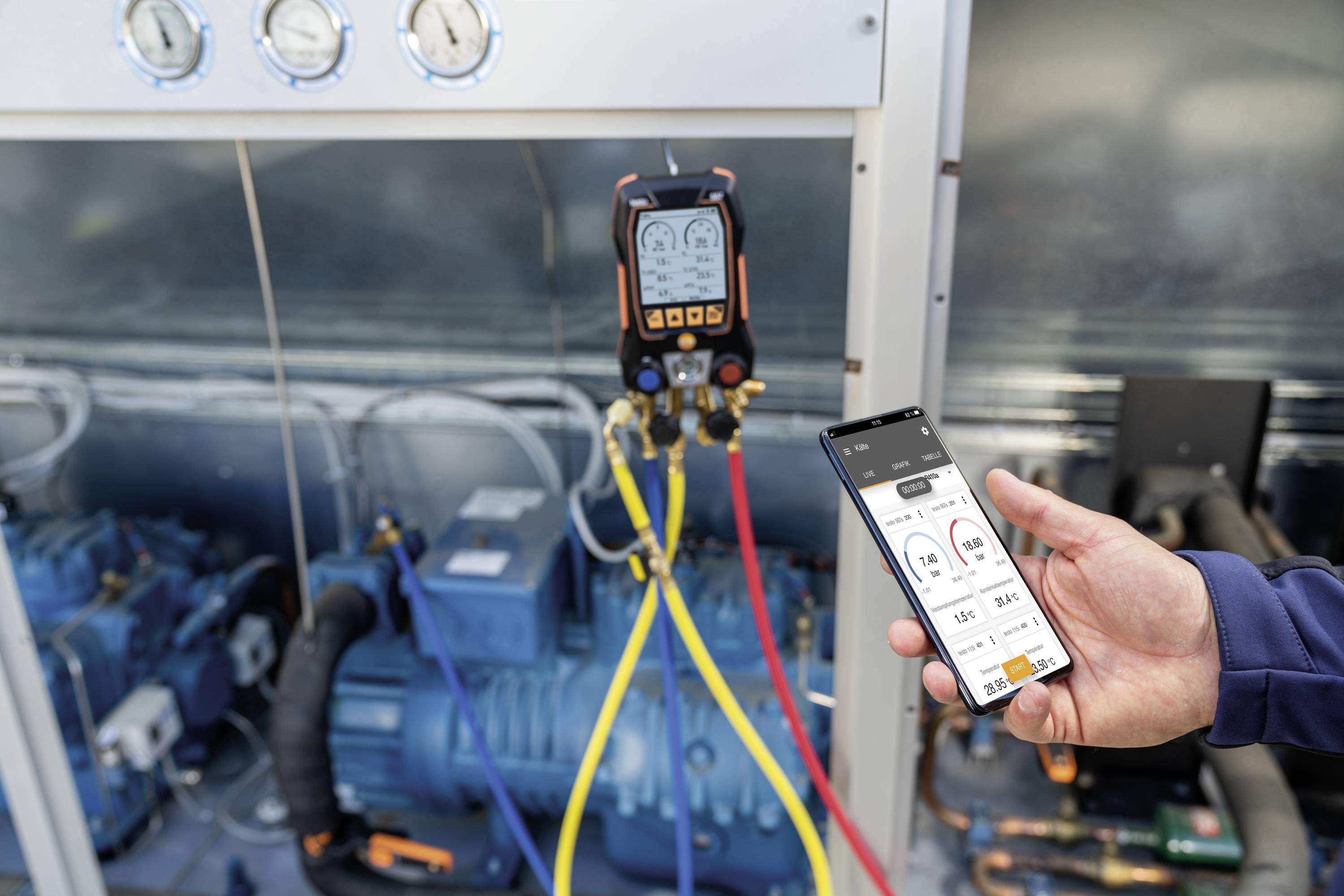 A person is holding a smartphone in front of a technical installation. Digital displays for system monitoring are visible on the screen.