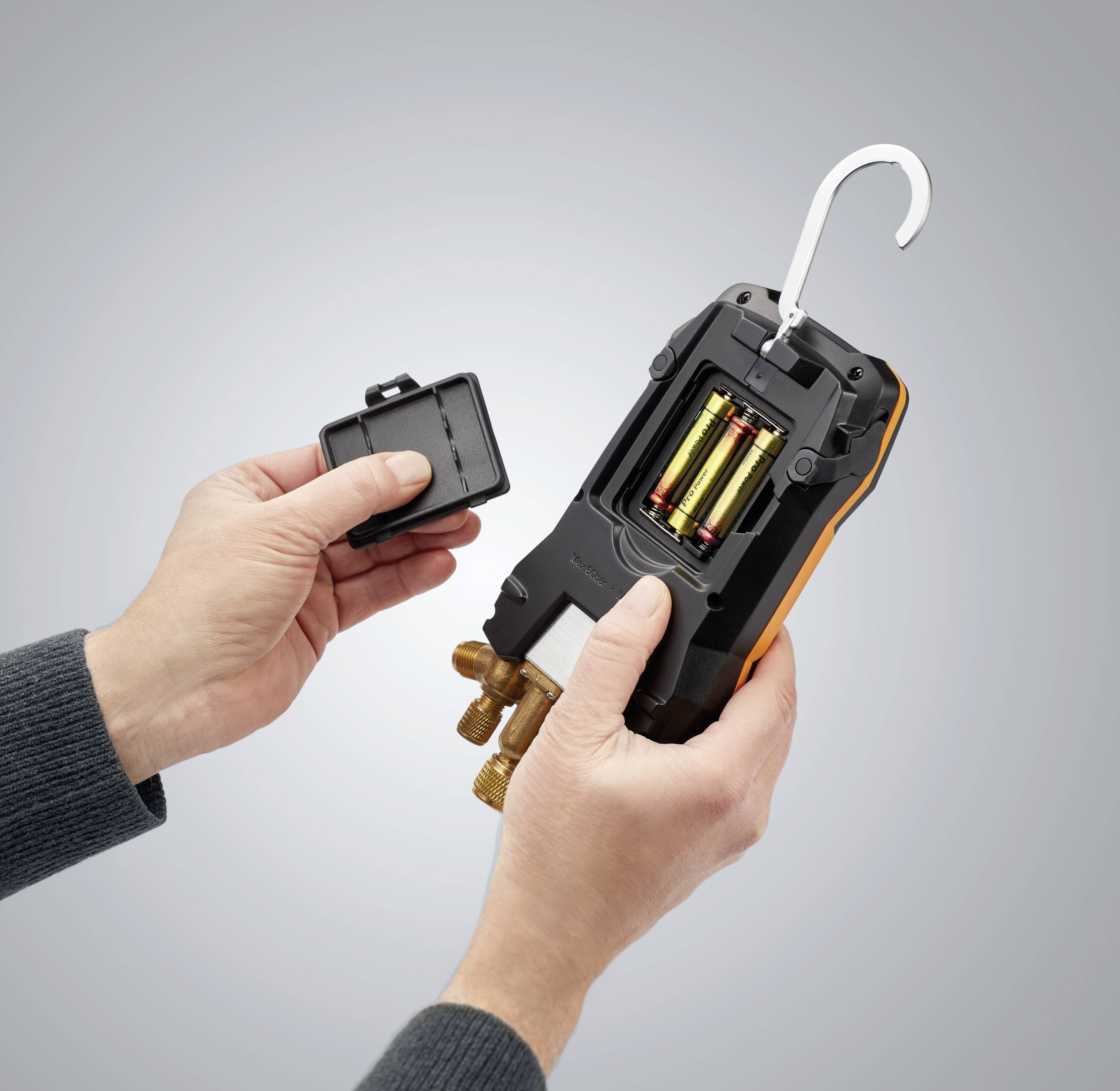 A person is holding a digital HVAC measuring device with the battery compartment cover open, displaying the inserted batteries.