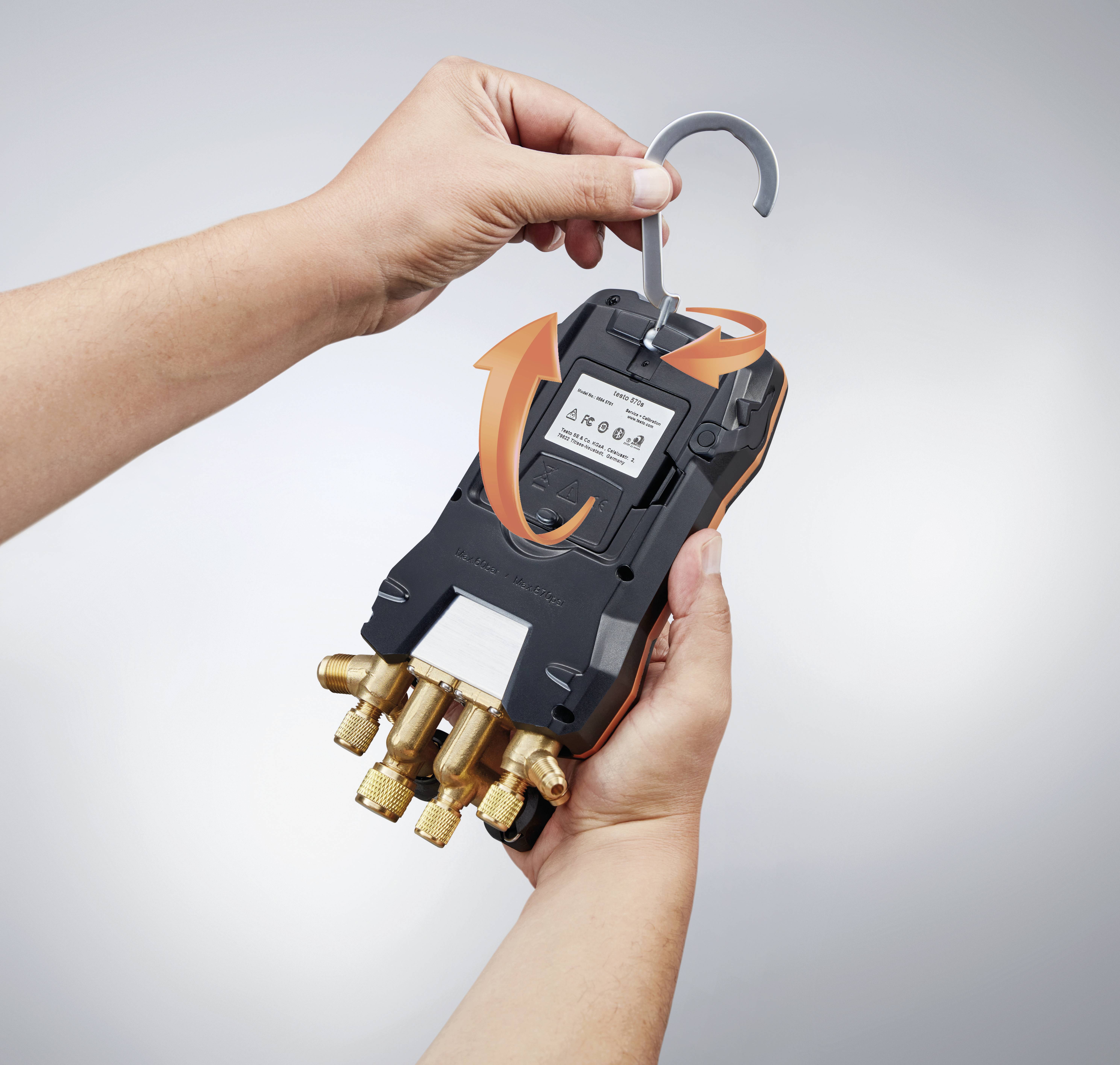 A hand is holding an electronic measuring device with a hook, while another hand points an arrow at the display. The device has multiple ports.