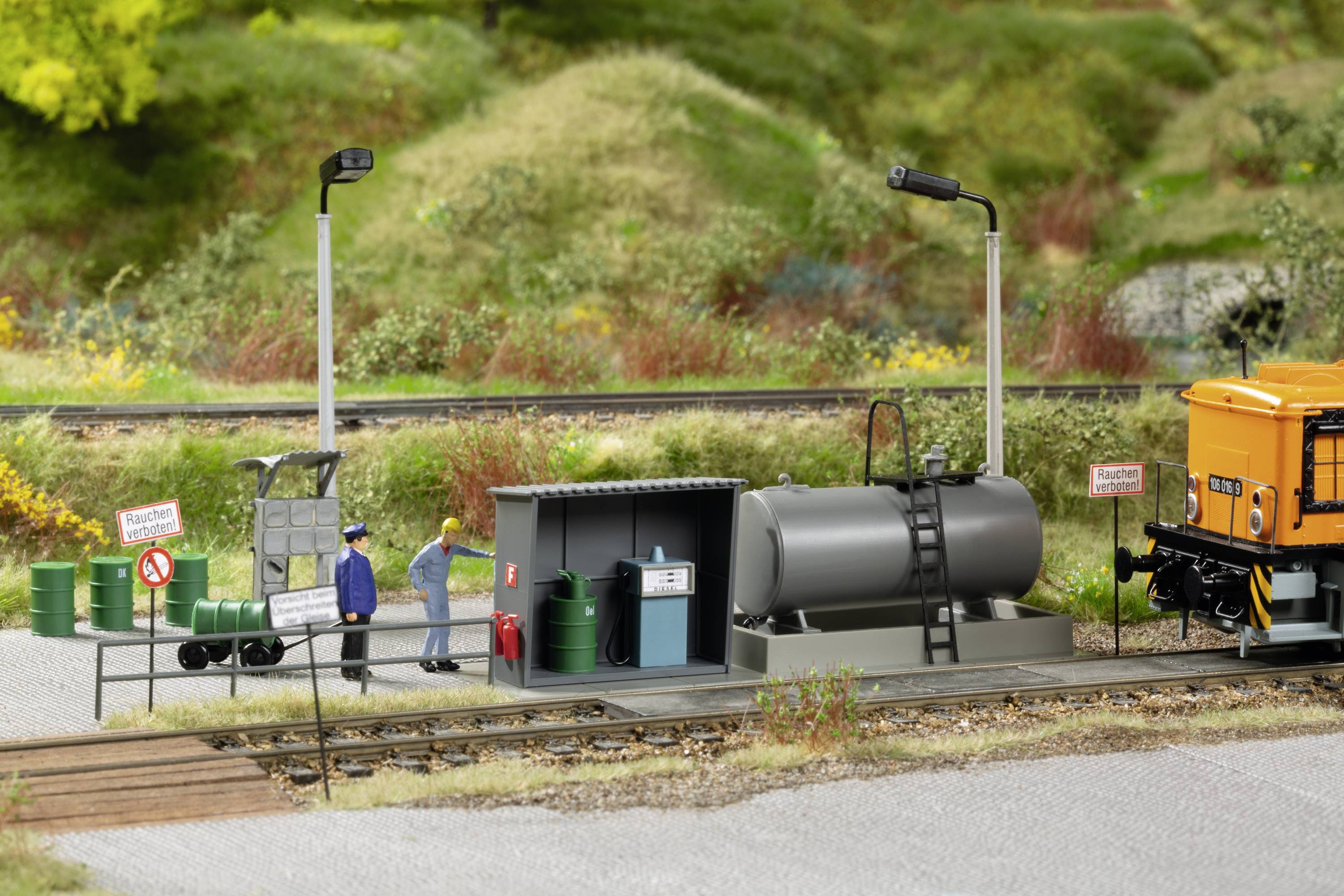 A small model of a railway refuelling station with two figures, a fuel tank, signs, and a yellow train beside a track, surrounded by green countryside.