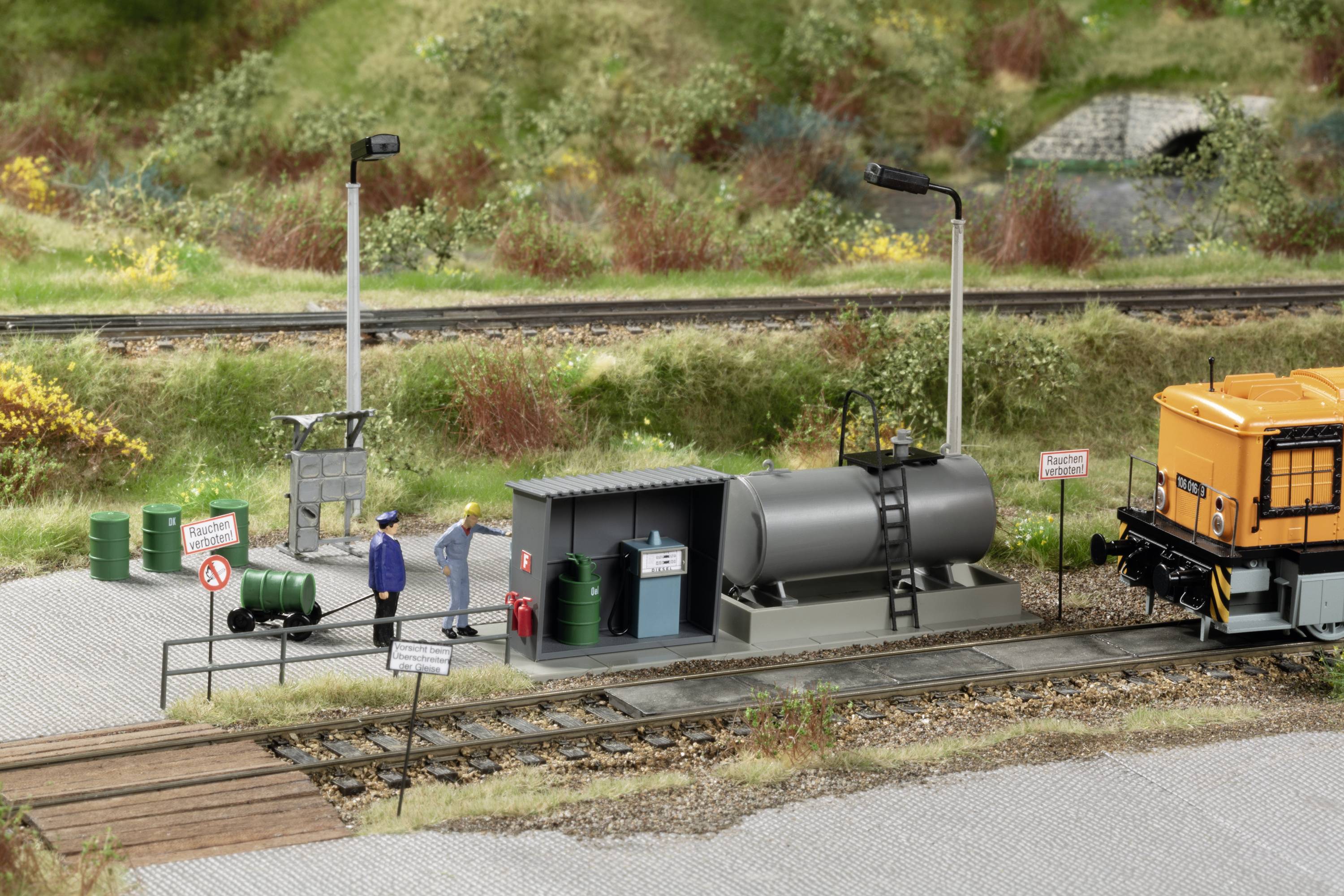 A small diorama depicts a car park with a refuelling station for locomotives. Two figures stand beside tanks. An orange vehicle is visible. Signs warn against unauthorised entry.