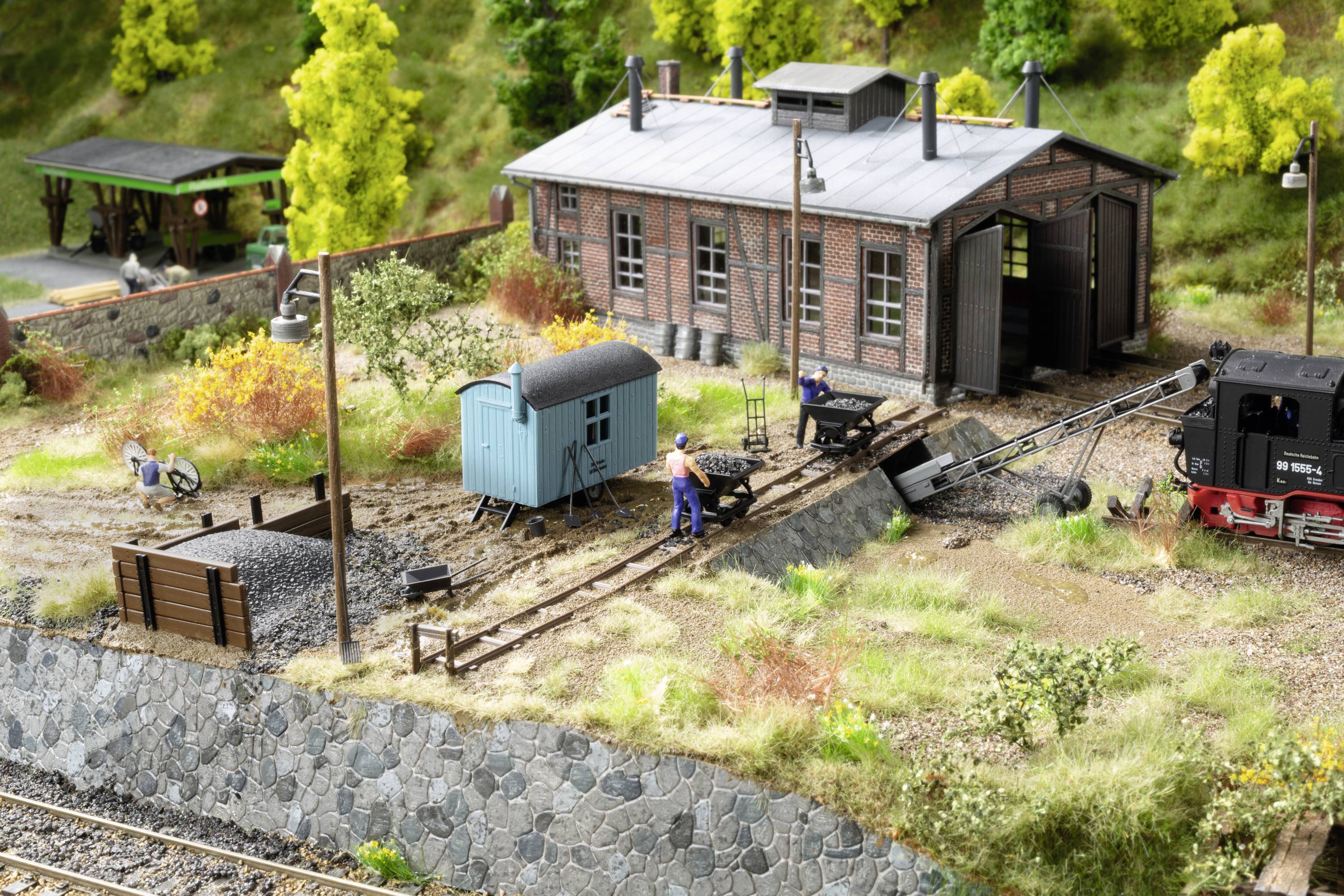 A miniature landscape model depicts a railway depot with a blue construction wagon, tracks, and people working on them, with trees in the background.