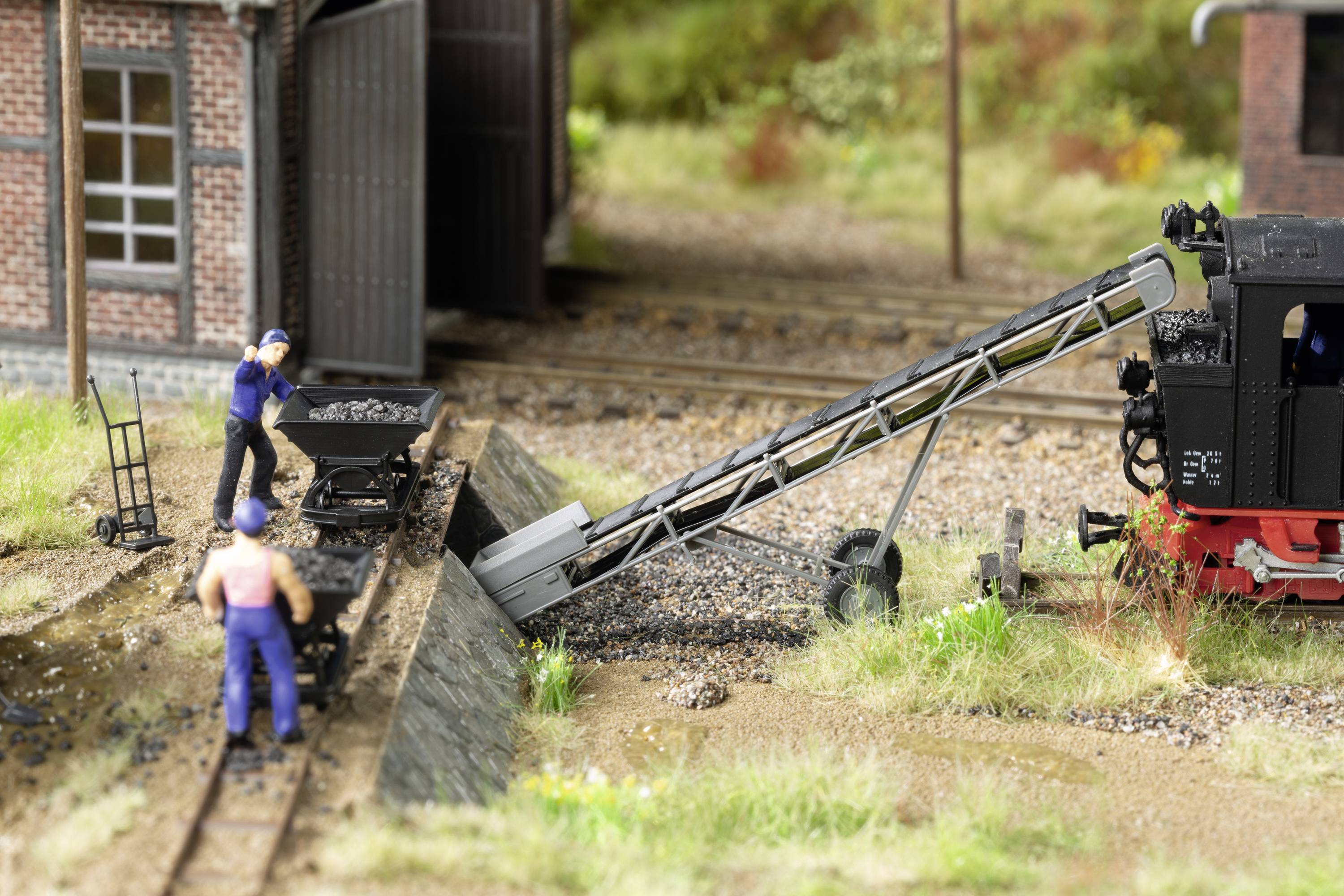 Two miniature figures are working on a model railway layout, using a conveyor belt to load a wagon with coal in front of a locomotive shed.