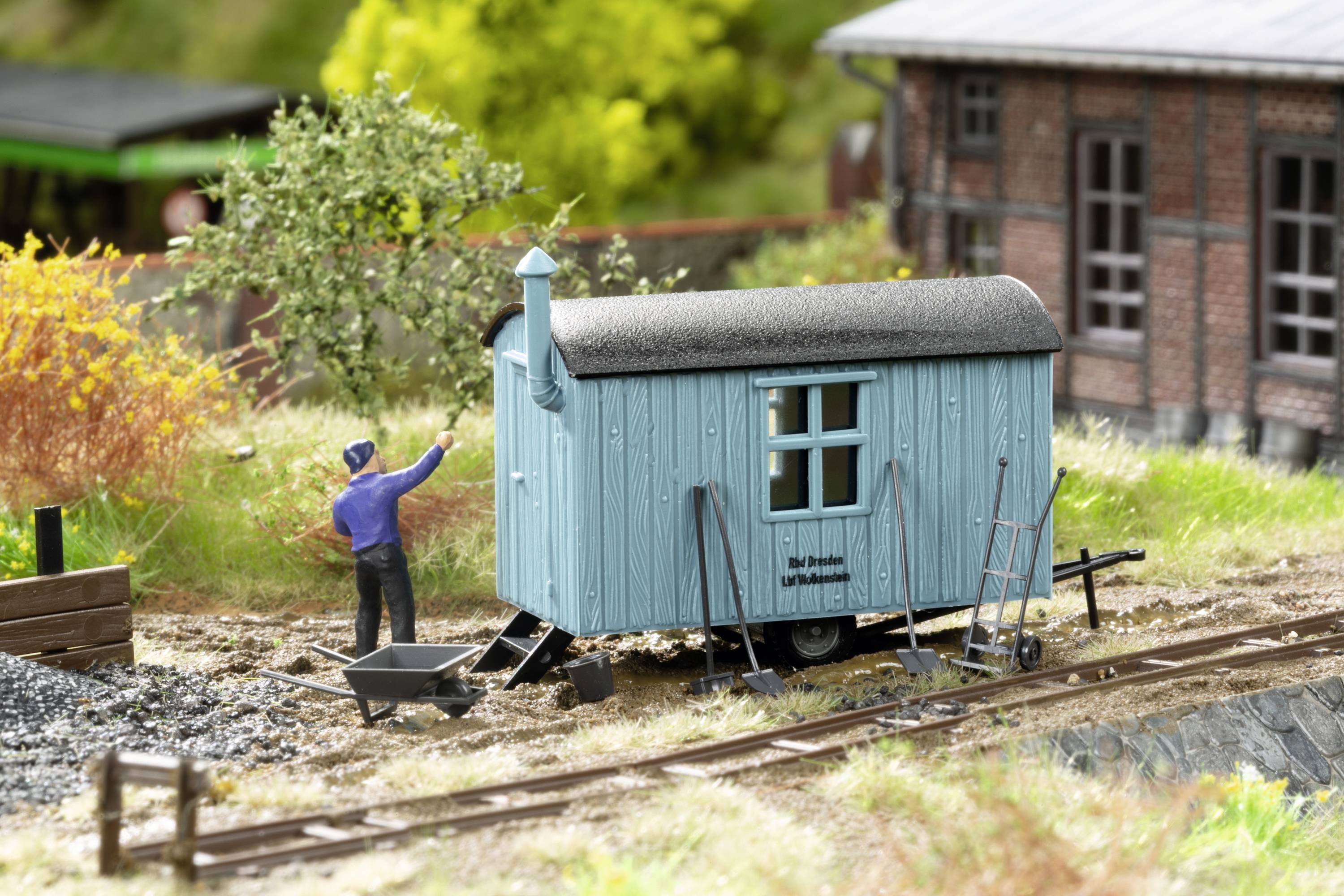 A model construction worker in blue clothing stands in front of a small blue construction trailer in a landscape with railway tracks and a brick building.