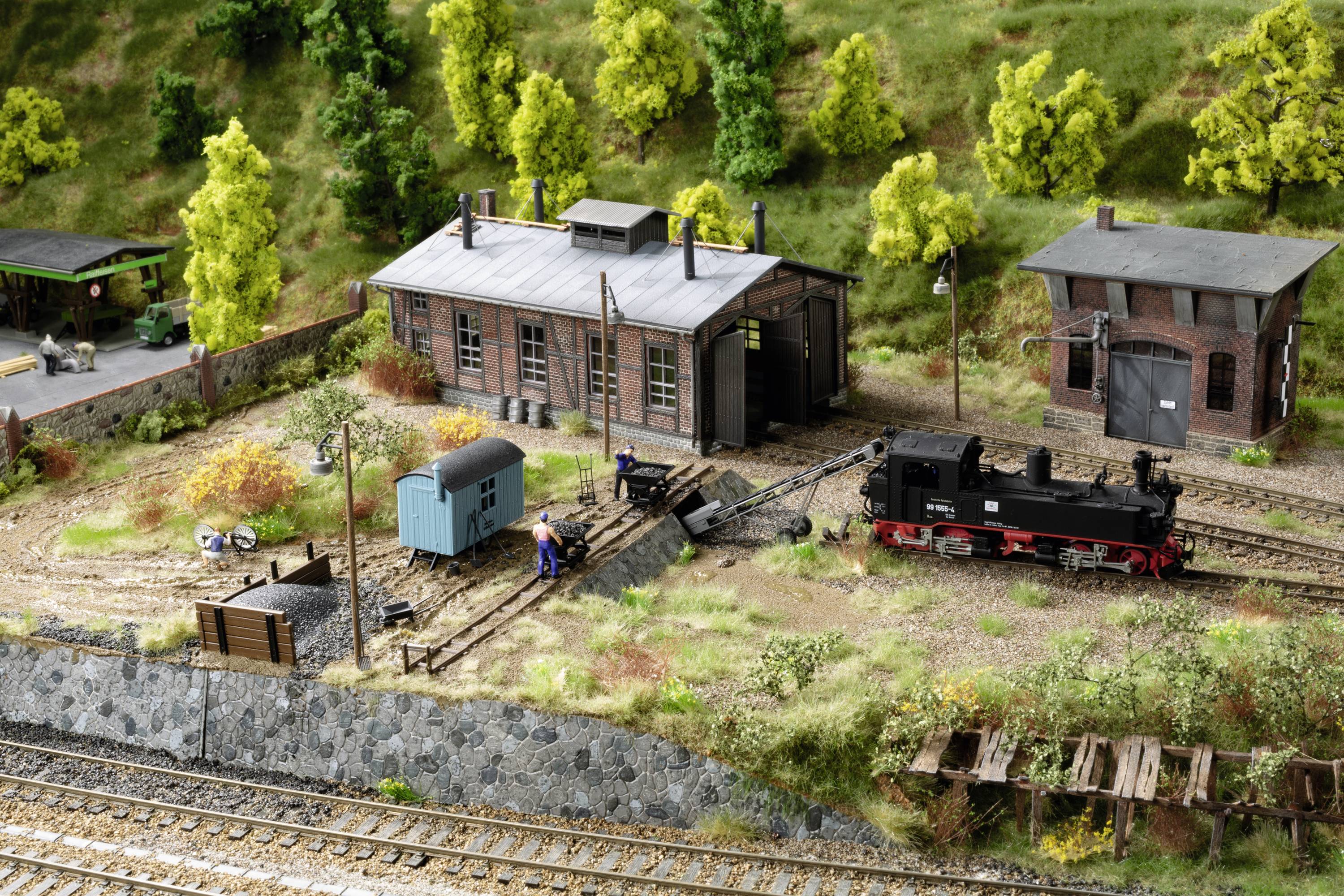 A miniature railway landscape with a small locomotive shed, tracks, a black steam locomotive and green forest in the background.