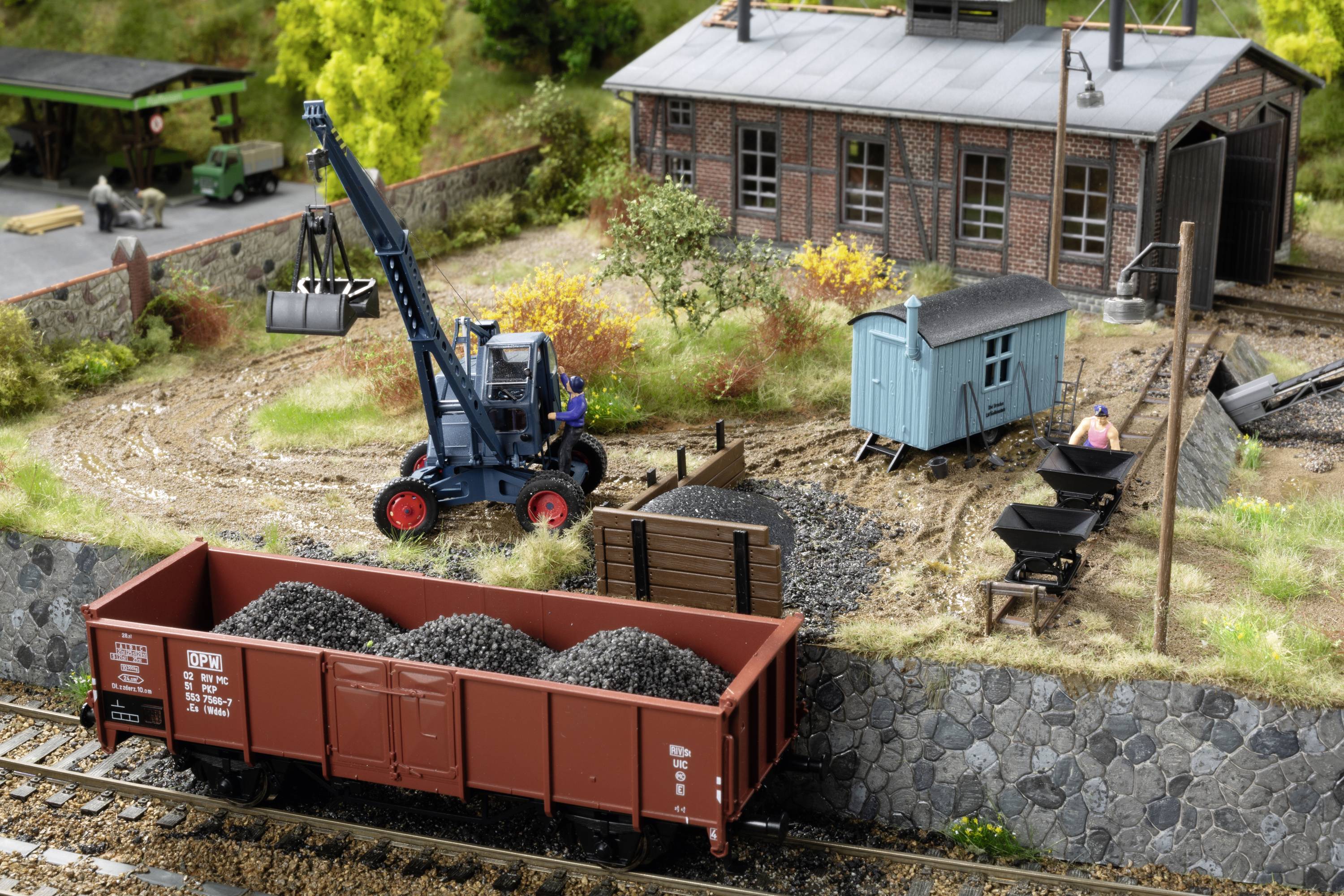 Model railway scene: A small excavator is lifting coal into a railway wagon, next to a red brick building and a blue shed.