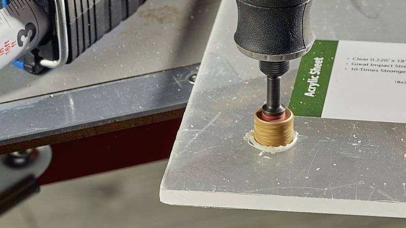 Close-up of a drill creating a hole in a perspex sheet. A label on the sheet indicates acrylic glass material.
