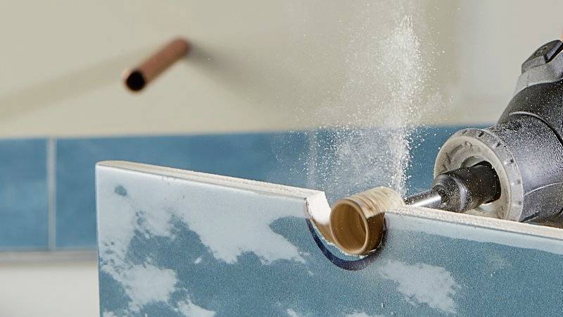 A drill pierces a tile in a wall. The tiles are blue and white patterned. Dust is created. Blurred details in the background.