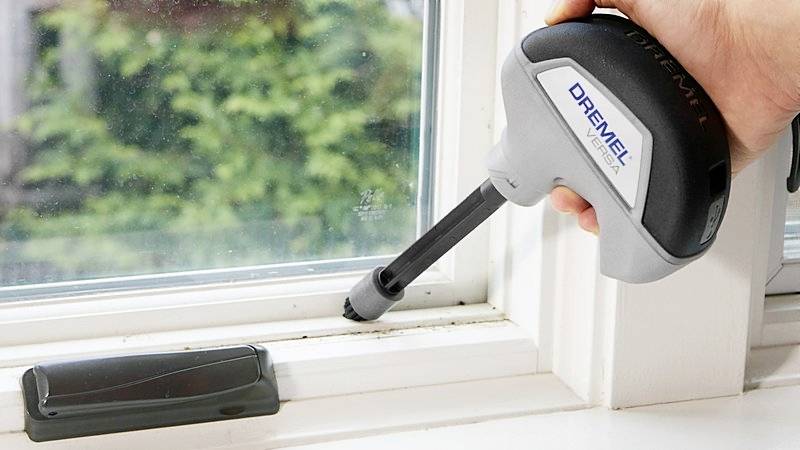 A person is cleaning dirt from a window frame using an electric tool, with a blurred window in the background.
