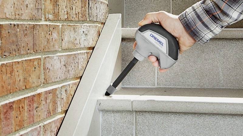 A person is cleaning the joints of a staircase next to a brick wall using a Dremel tool.