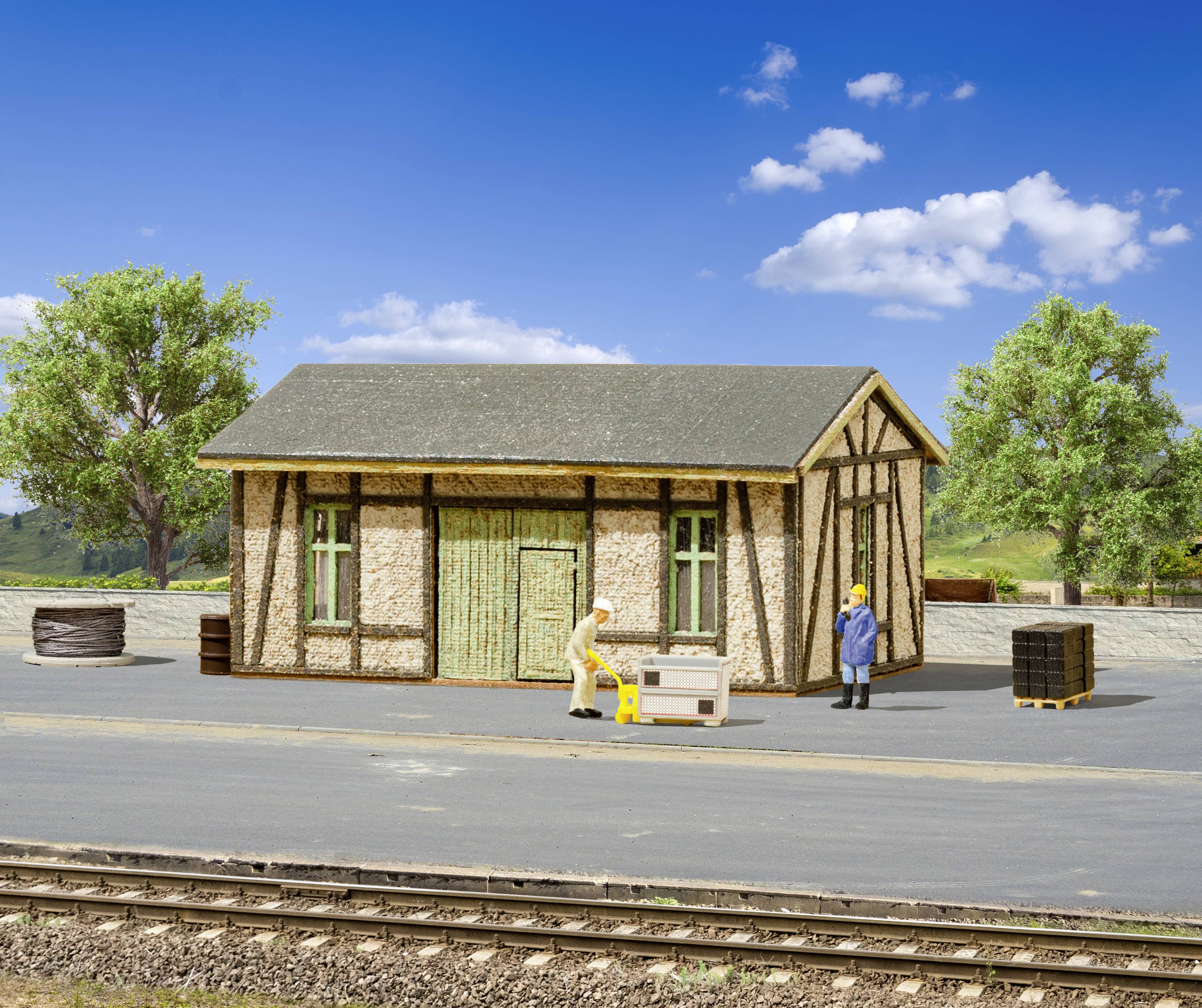 Small railway station building with two people loading crates. Surrounded by trees and blue sky. Tracks in the foreground.