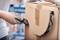 A person opens a cardboard box with a Stanley knife. They are wearing gloves. Blurred storage shelves are visible in the background.