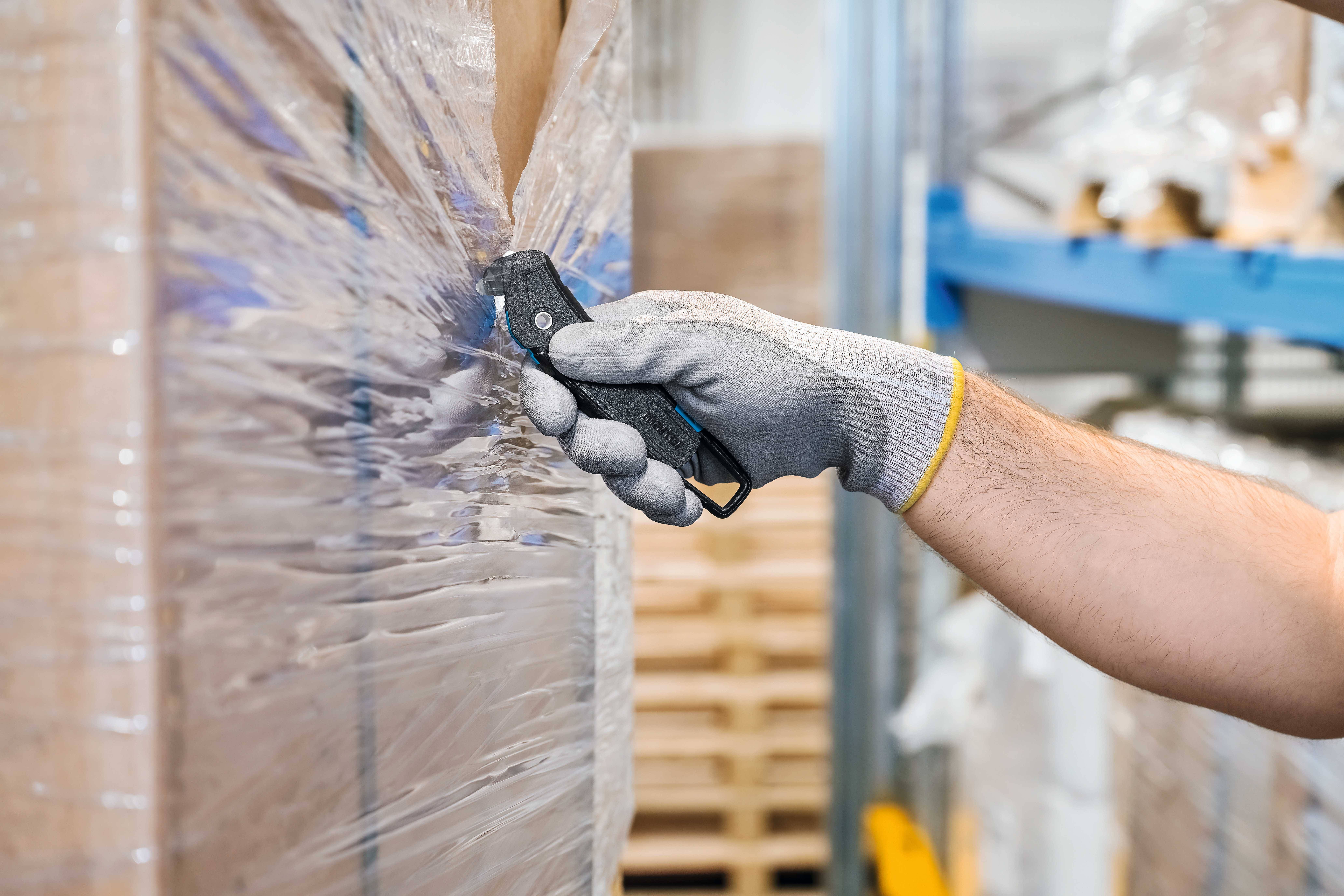 A person opens a pallet with a black security knife in a warehouse. They are wearing grey protective gloves.