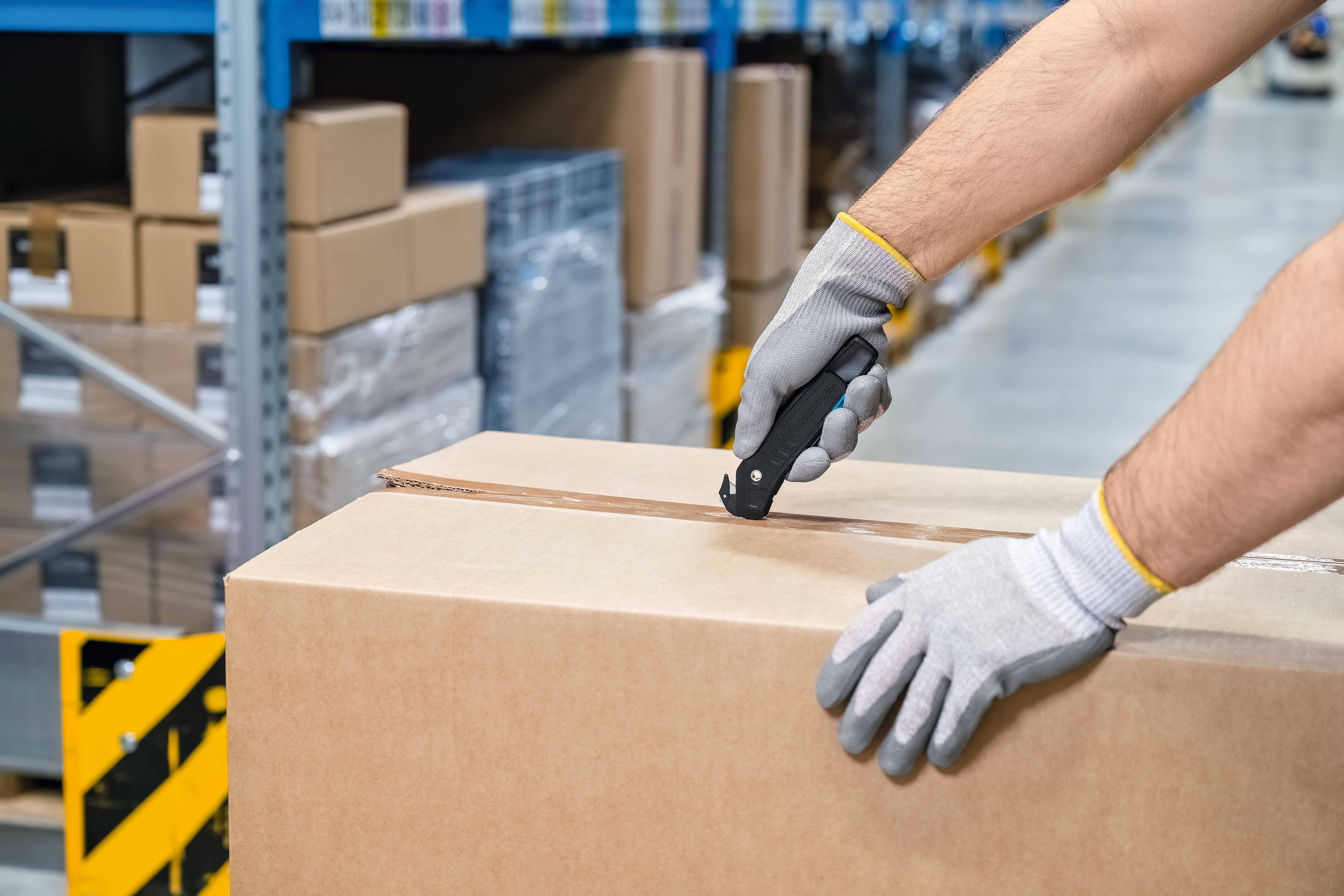 A person is opening a large cardboard box with a utility knife in a warehouse. Shelves with packages are visible in the background.