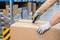A person is opening a large cardboard box with a utility knife in a warehouse. Shelves with packages are visible in the background.