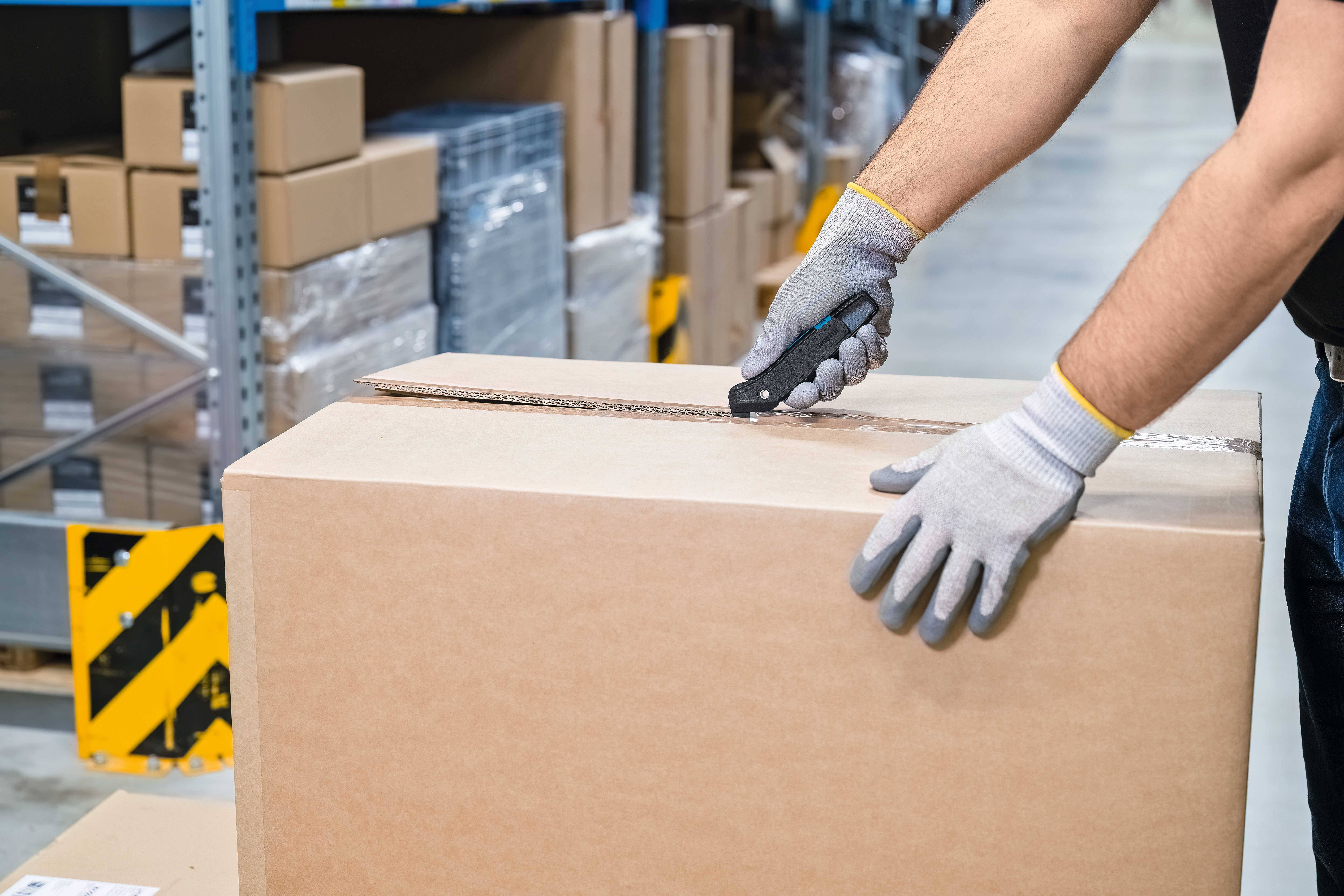 A person is opening a large package with a Stanley knife in a warehouse. Shelves with cardboard boxes are visible in the background.