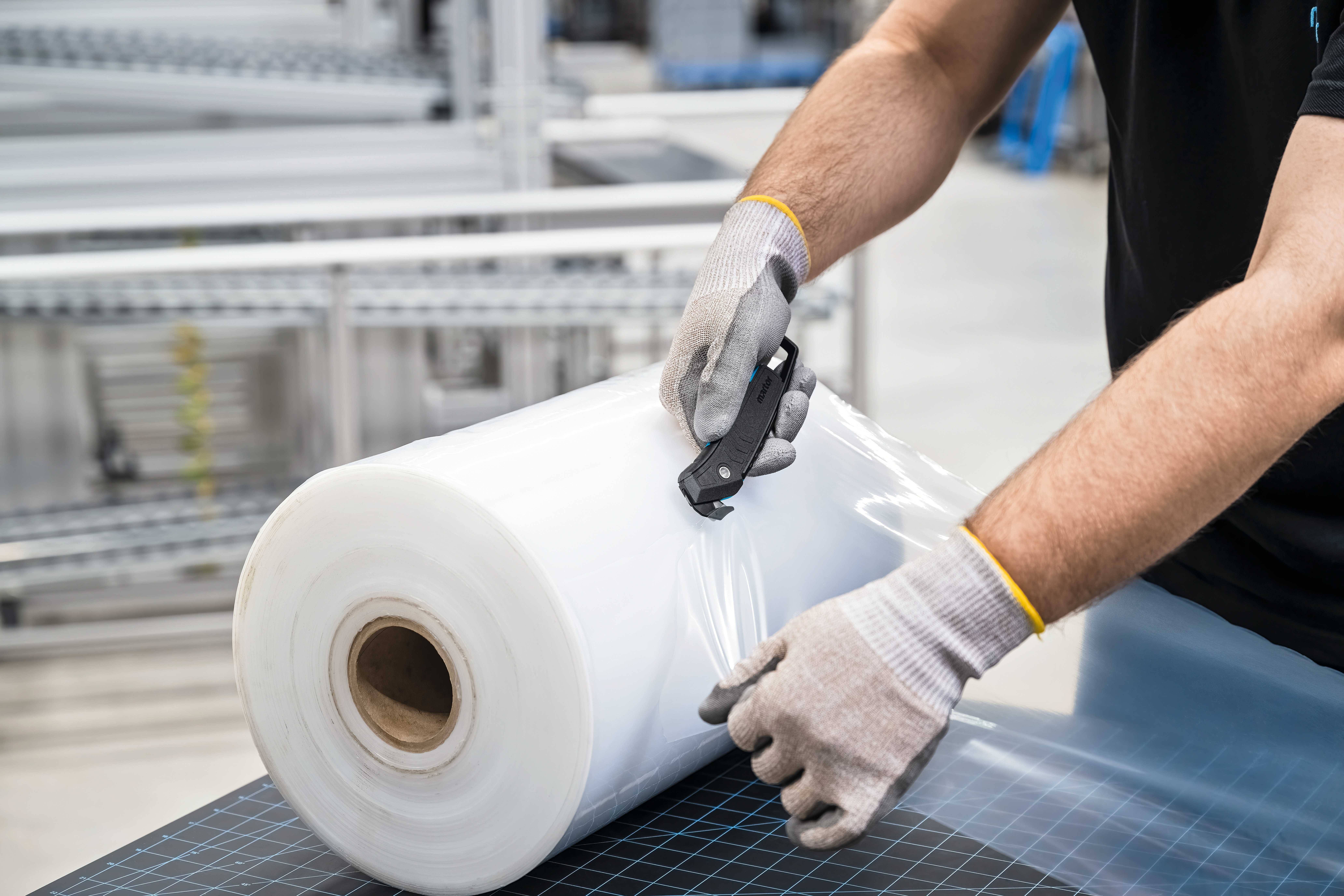A person is cutting plastic film from a large roll with a knife. Industrial shelving is visible in the background.