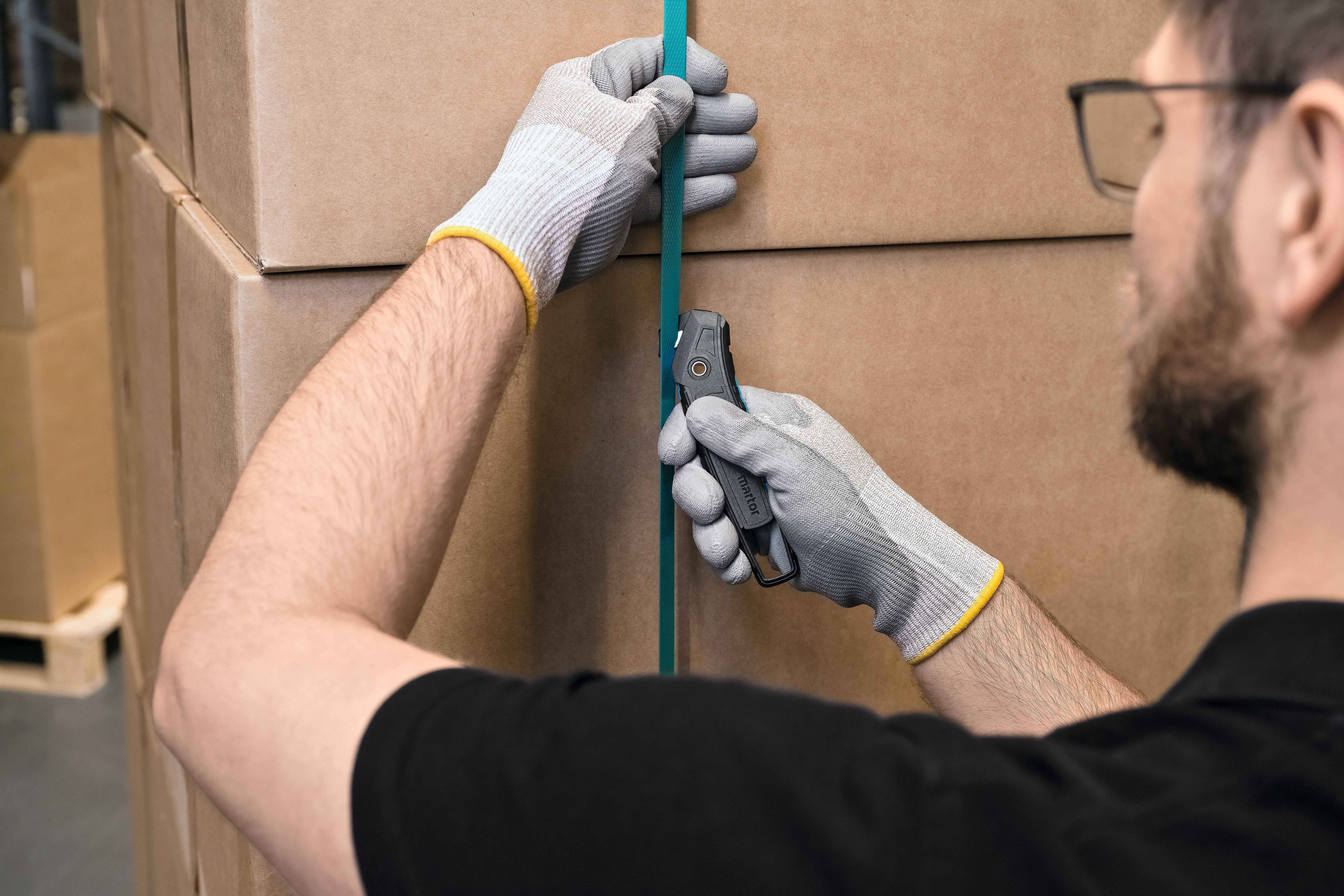 A person wearing gloves is cutting a green ribbon from a cardboard stack using a safety knife in a warehouse.
