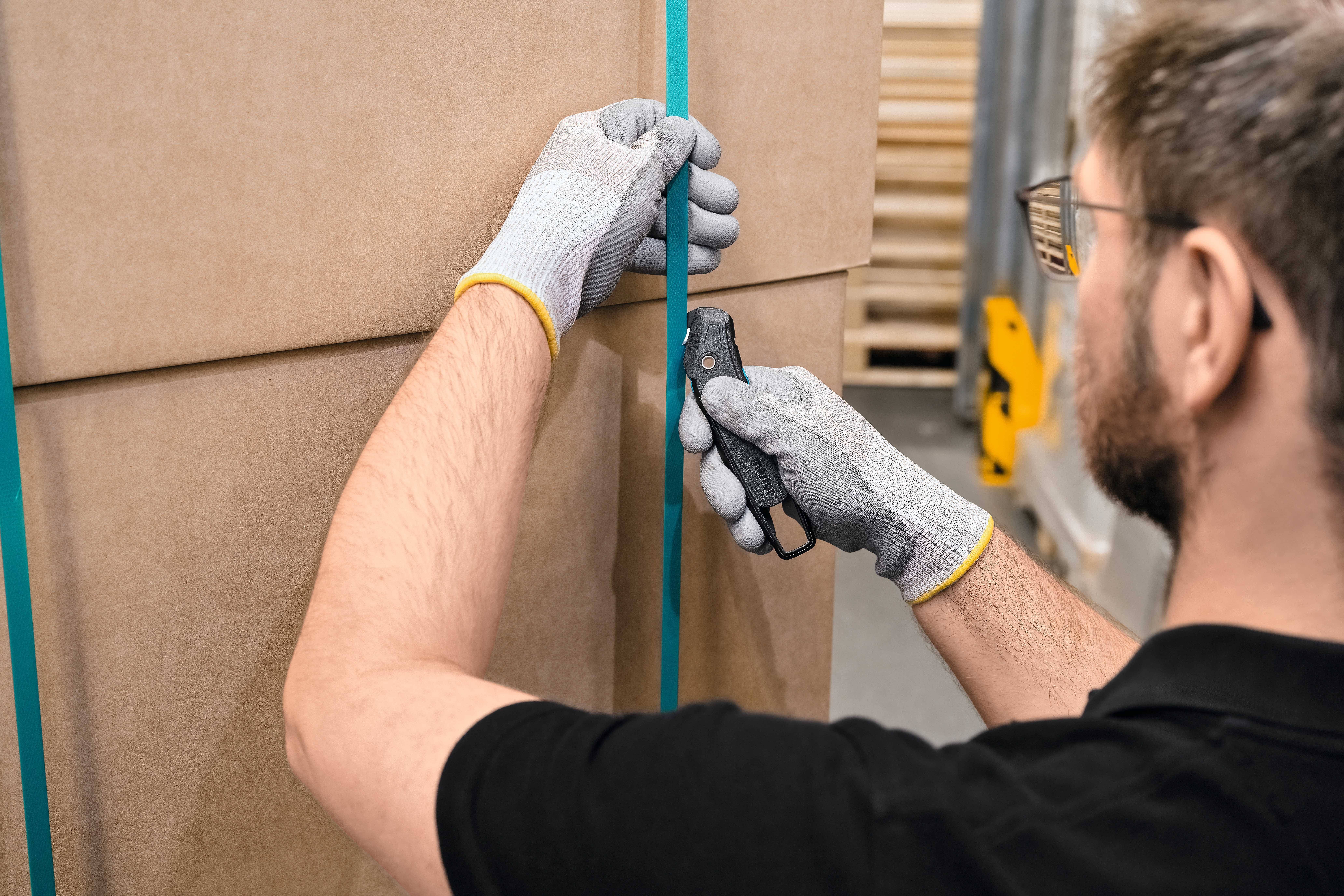 A man wearing gloves opens a cardboard box with a knife. The packaging is sealed with blue tape.