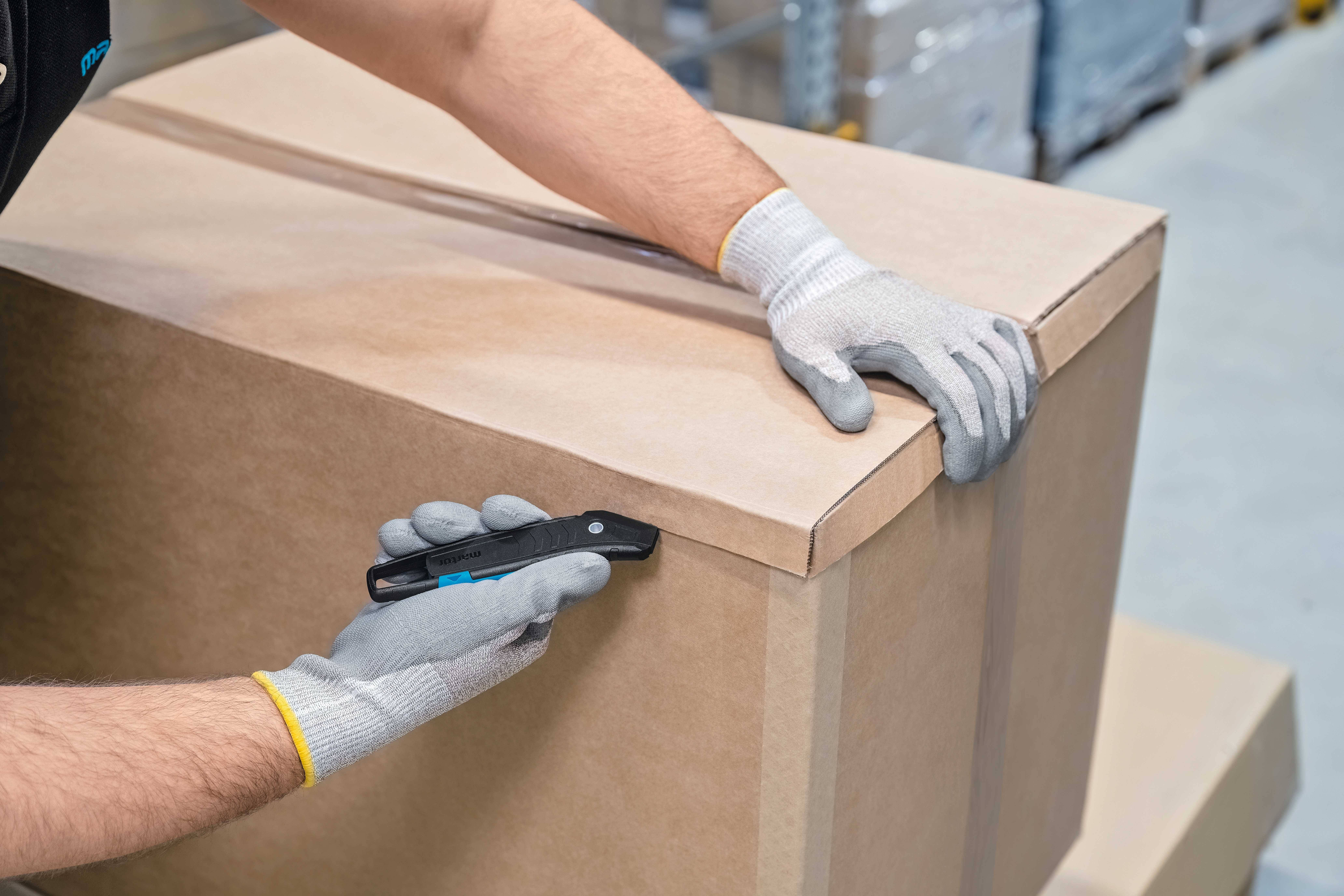A person wearing gloves opens a large cardboard box with a utility knife in a warehouse.